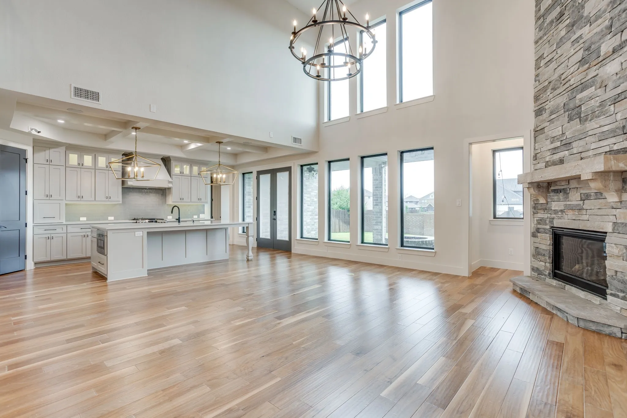 Spacious open-concept living area featuring wood-finish flooring, a stone-clad fireplace with a hearth, and a double-height ceiling with a prominent chandelier