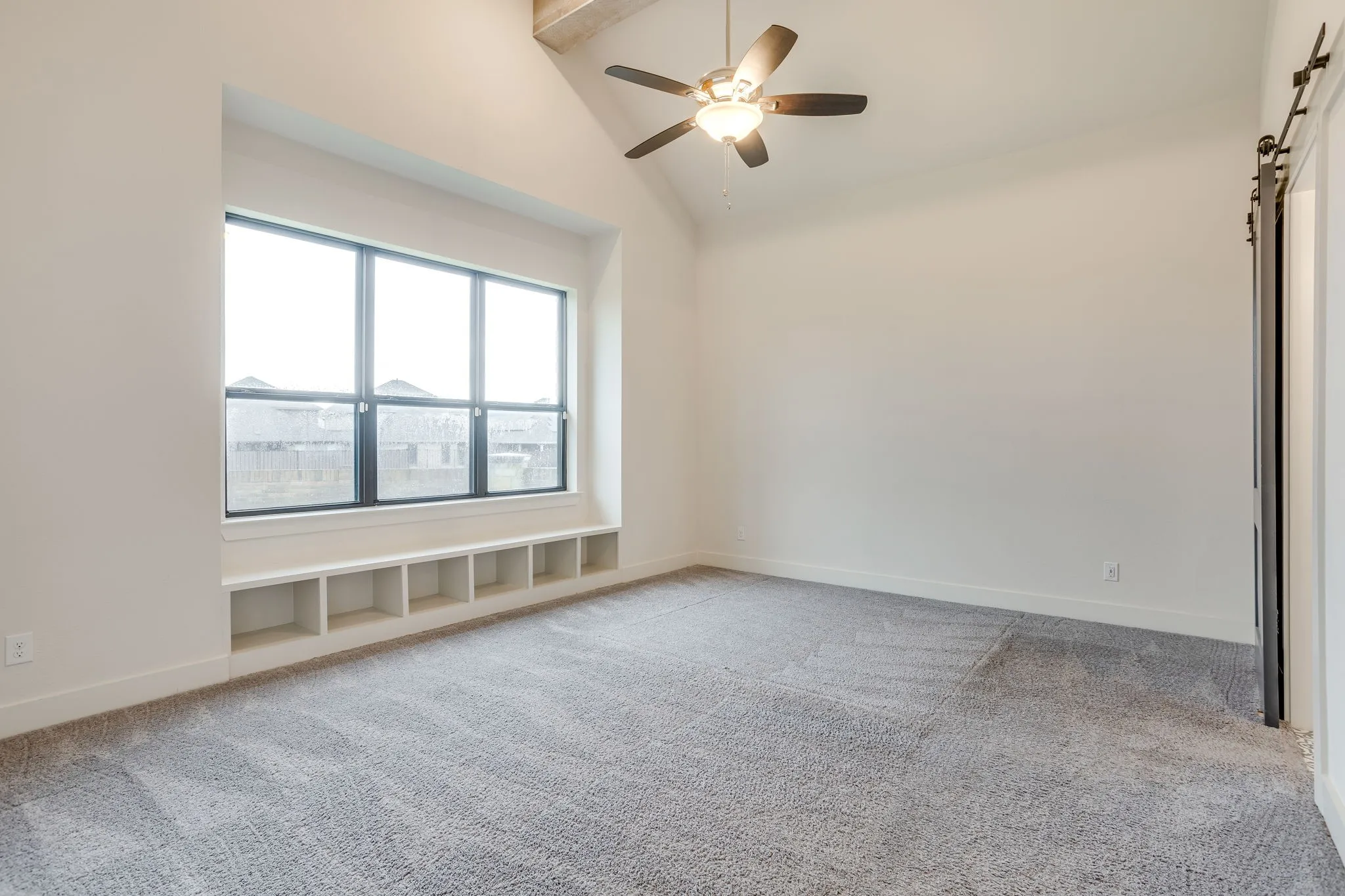 Spacious room featuring a large window with an integrated built-in storage bench, vaulted ceiling, a ceiling fan with integrated lighting, and wall-to-wall carpeting