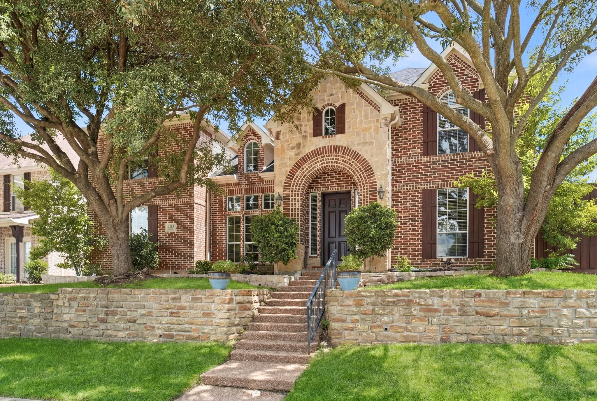 Stunning stone and brick exterior elevation featuring a grand brick-arched entryway, beautifully complemented by two tall, mature trees in the front yard that provide both aesthetic curb appeal and refreshing natural shade.