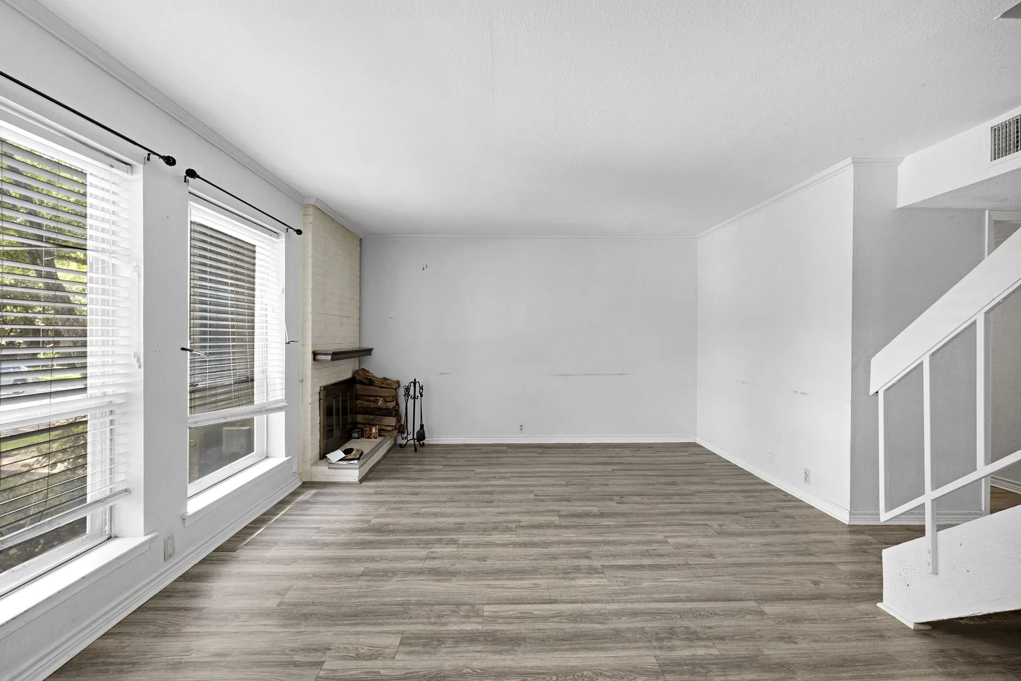 Spacious living area featuring wood-finish flooring, a white brick fireplace with a dark wood mantel, and multiple windows with white blinds