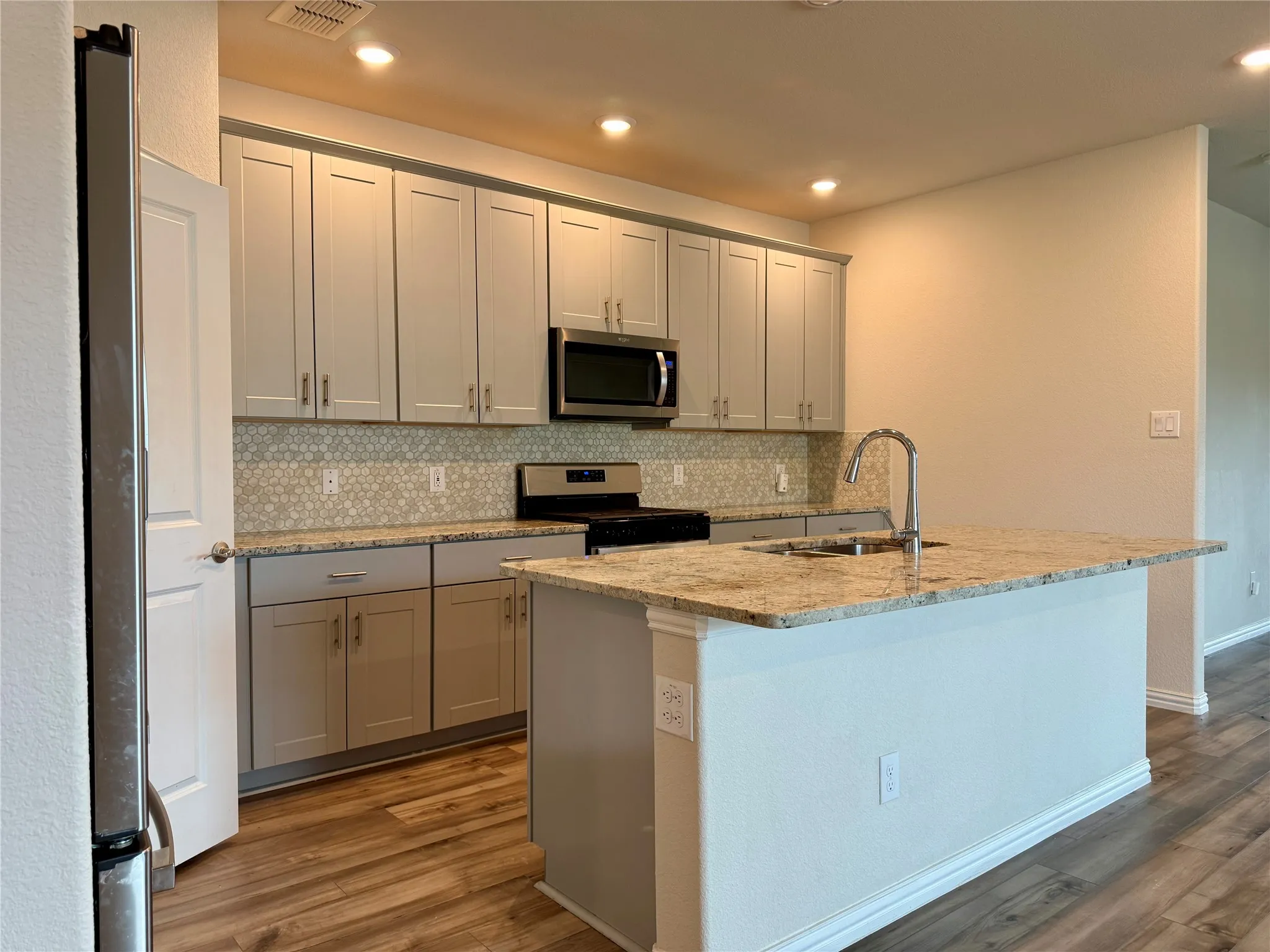 Kitchen featuring wood-finish flooring, light grey cabinetry, stainless steel appliances, and a central island with a granite countertop and integrated sink