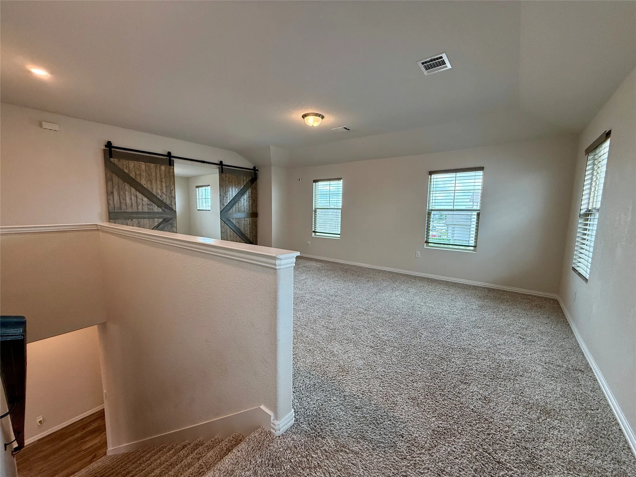 Spacious upper-level room featuring barn door entry, plush carpet flooring, multiple windows, recessed lighting, and a neutral color palette
