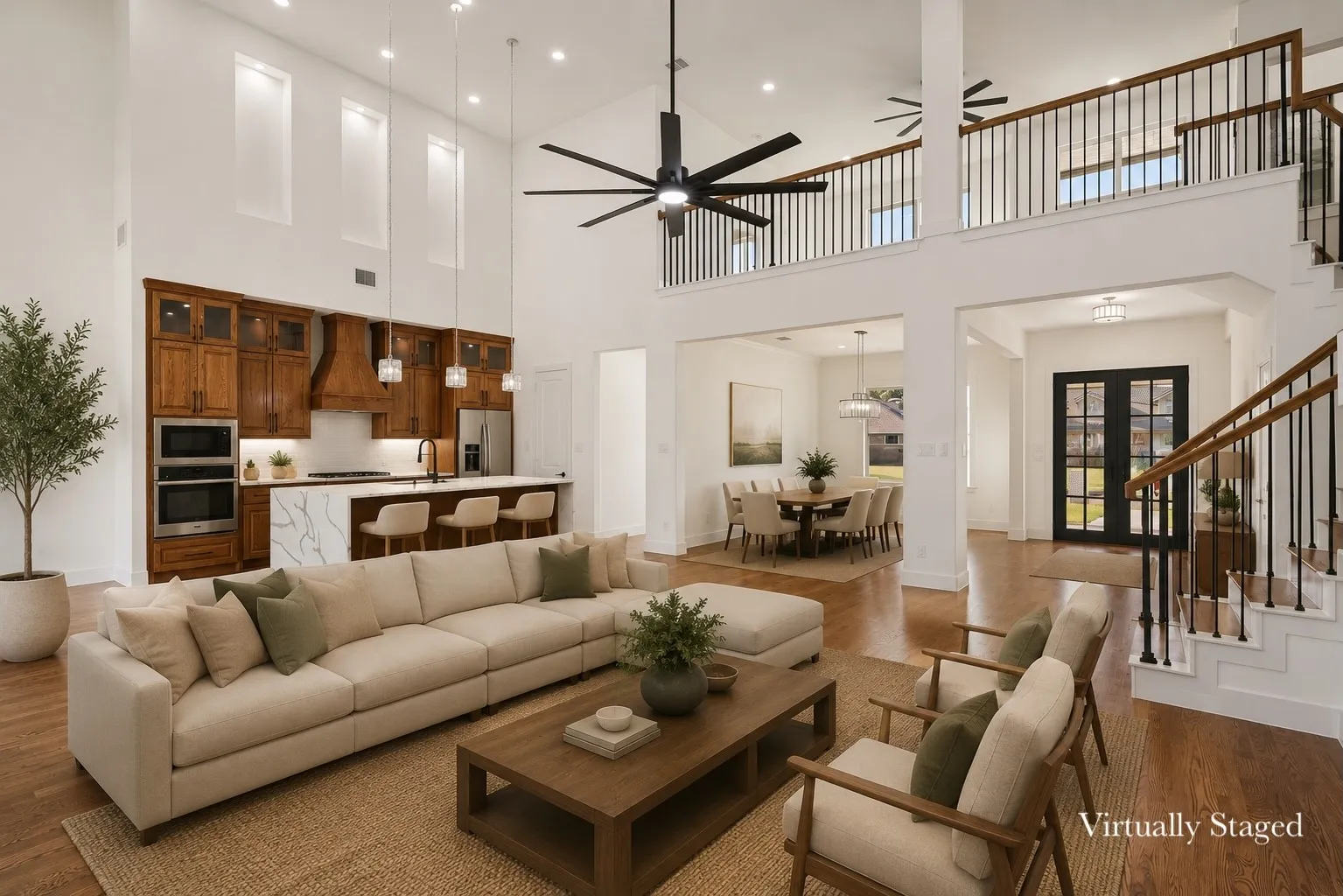 Open-concept living space featuring high ceilings with recessed lighting, wood-finish flooring, a kitchen with rich wood cabinetry and a large island, stainless steel double ovens, and an open second-story walkway with a black iron railing