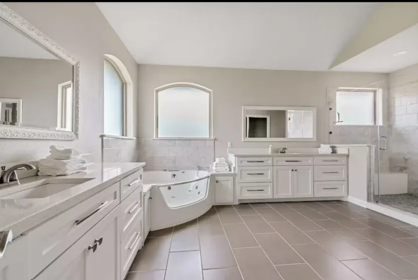 Spacious bathroom featuring a corner soaking tub, dual vanities with white cabinetry, a glass-enclosed shower, and large format tile flooring
