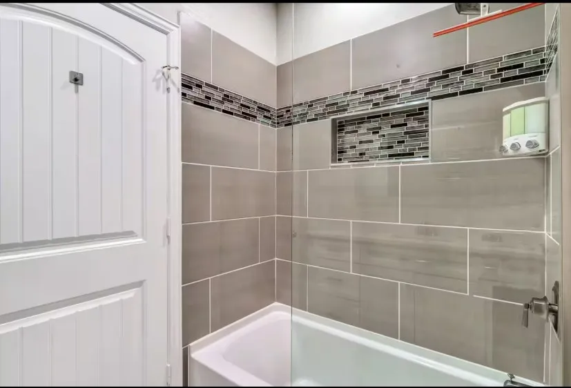Bathroom featuring a white tub, large format gray wall tiling, a decorative glass mosaic tile accent band, a recessed niche with mosaic tiling, and satin nickel plumbing fixtures