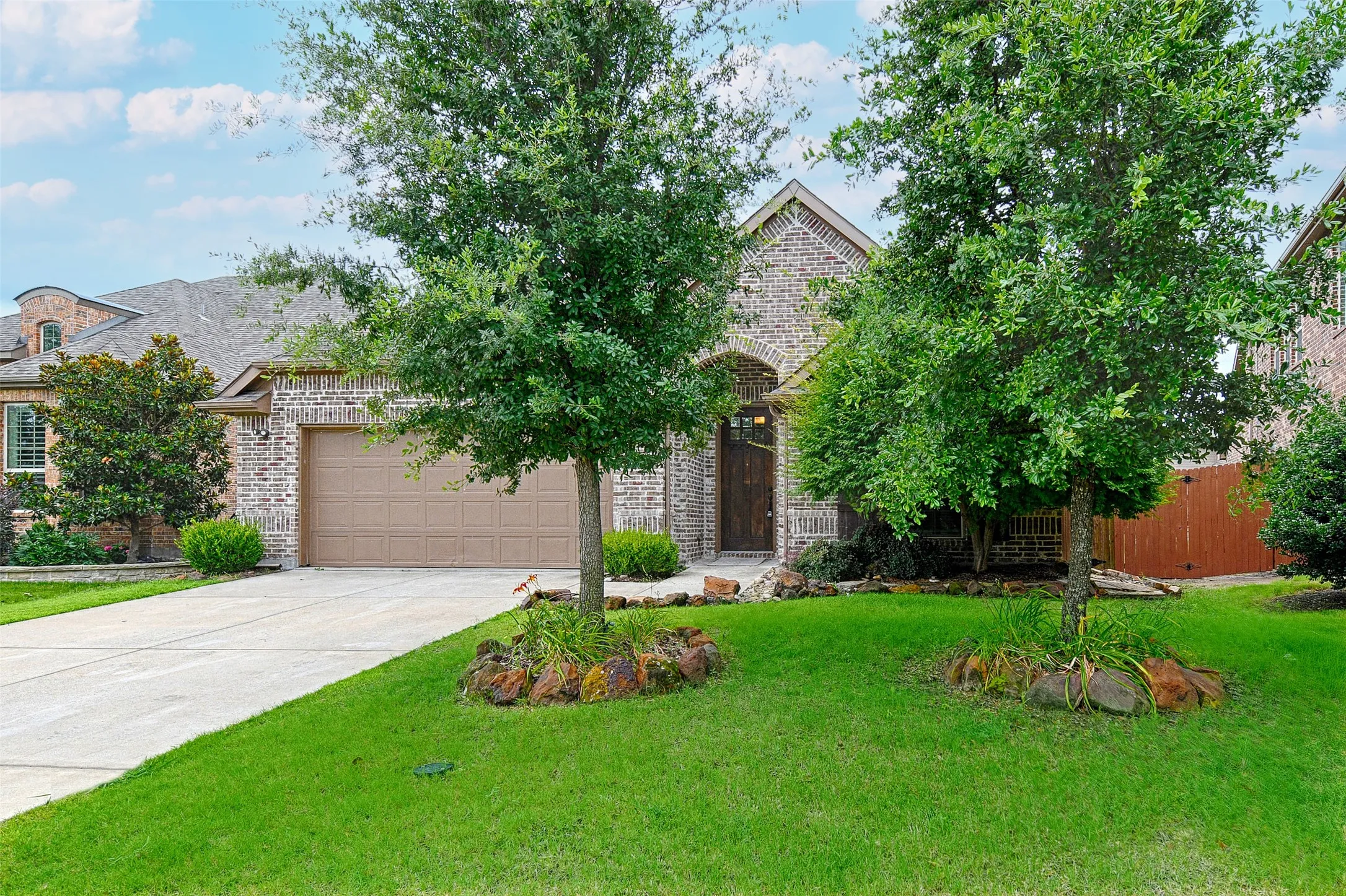 Brick exterior featuring an arched entryway, integrated two-car garage, concrete driveway, and established front landscaping with mature trees