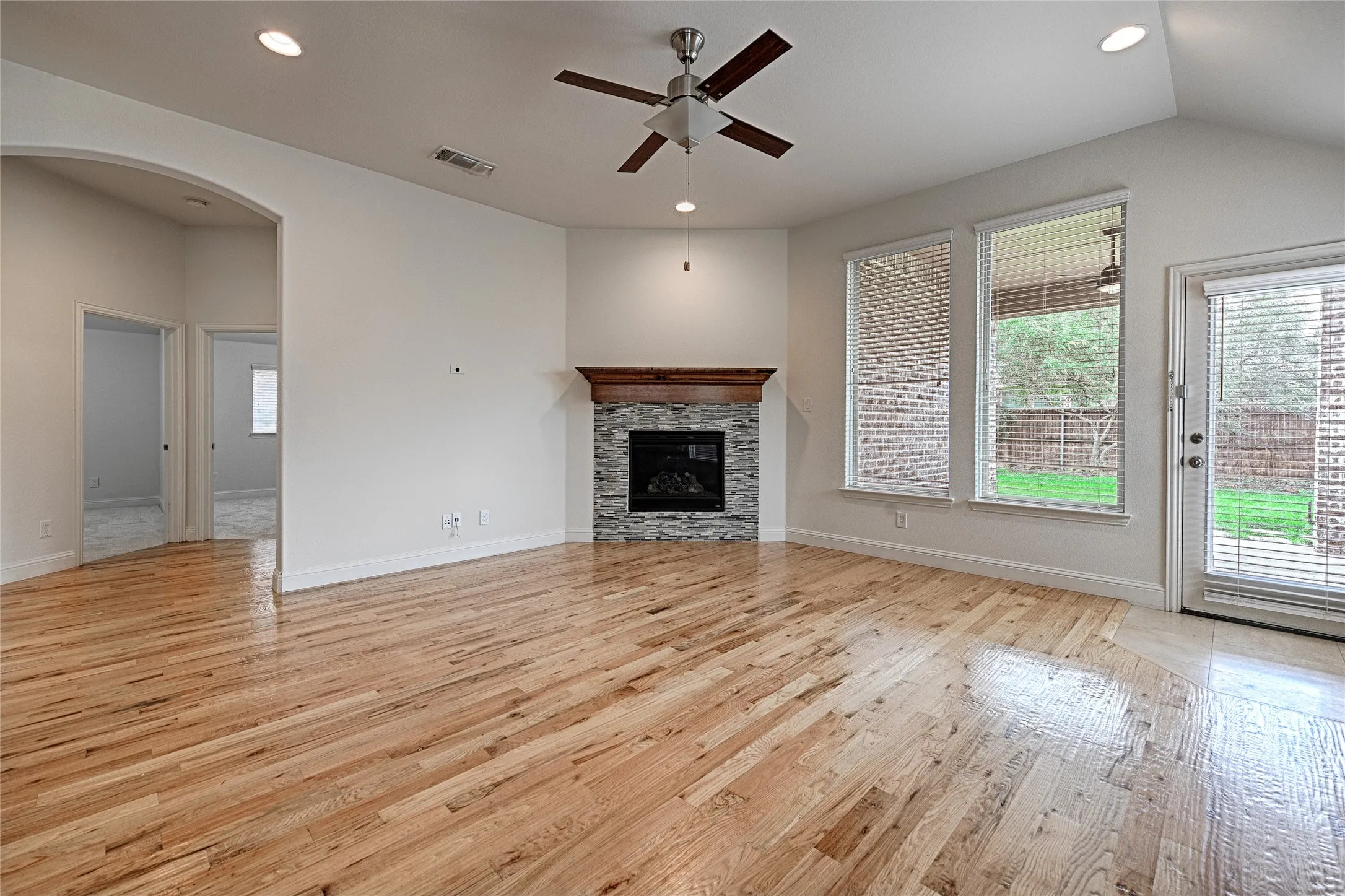 Spacious interior featuring wood-finish flooring, a stone-front fireplace with wood mantel, a ceiling fan, recessed lighting, and multiple windows with blinds