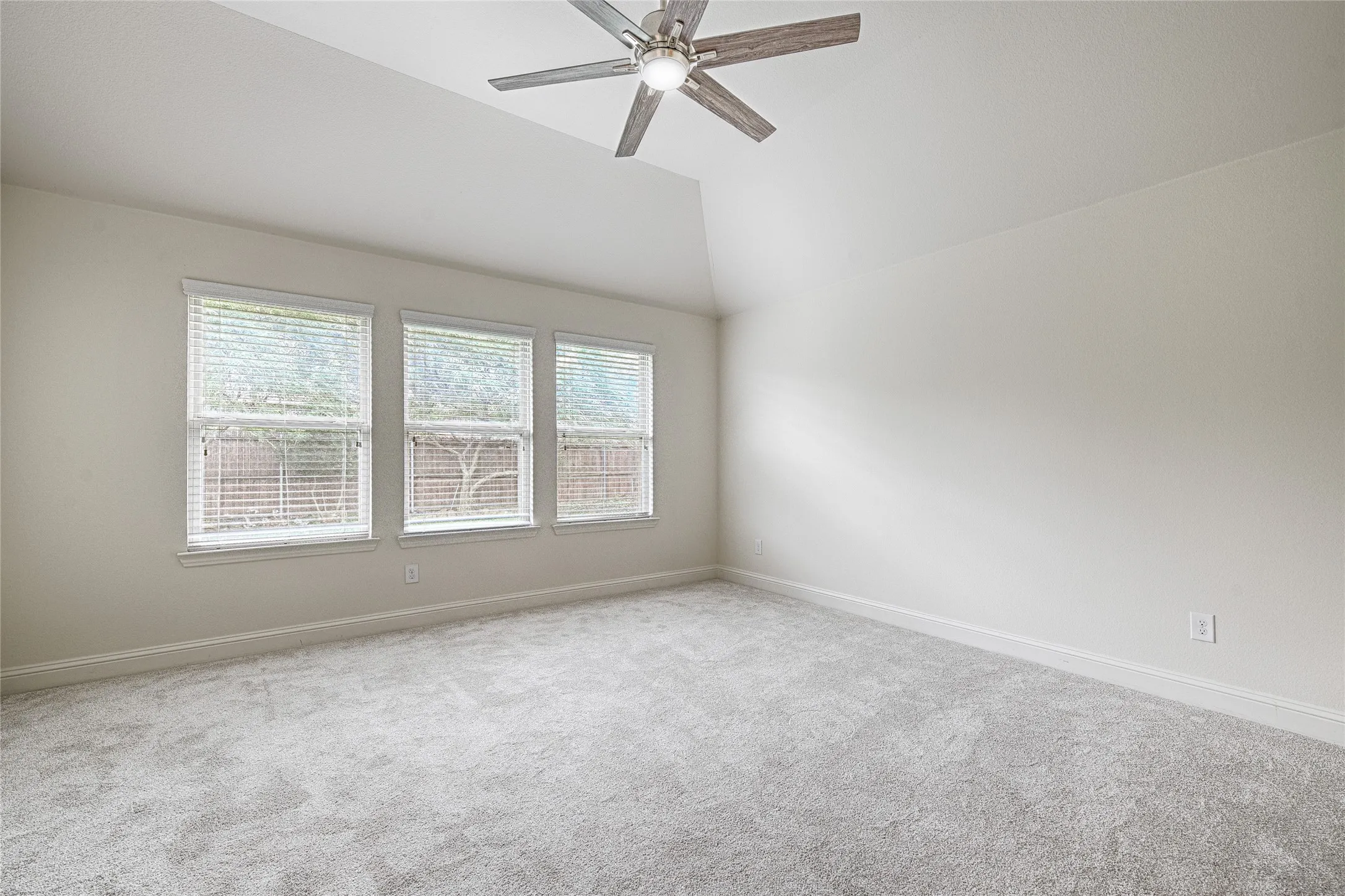 Spacious room featuring vaulted ceilings, light gray carpeting, three large windows with blinds, and a contemporary ceiling fan with integrated lighting