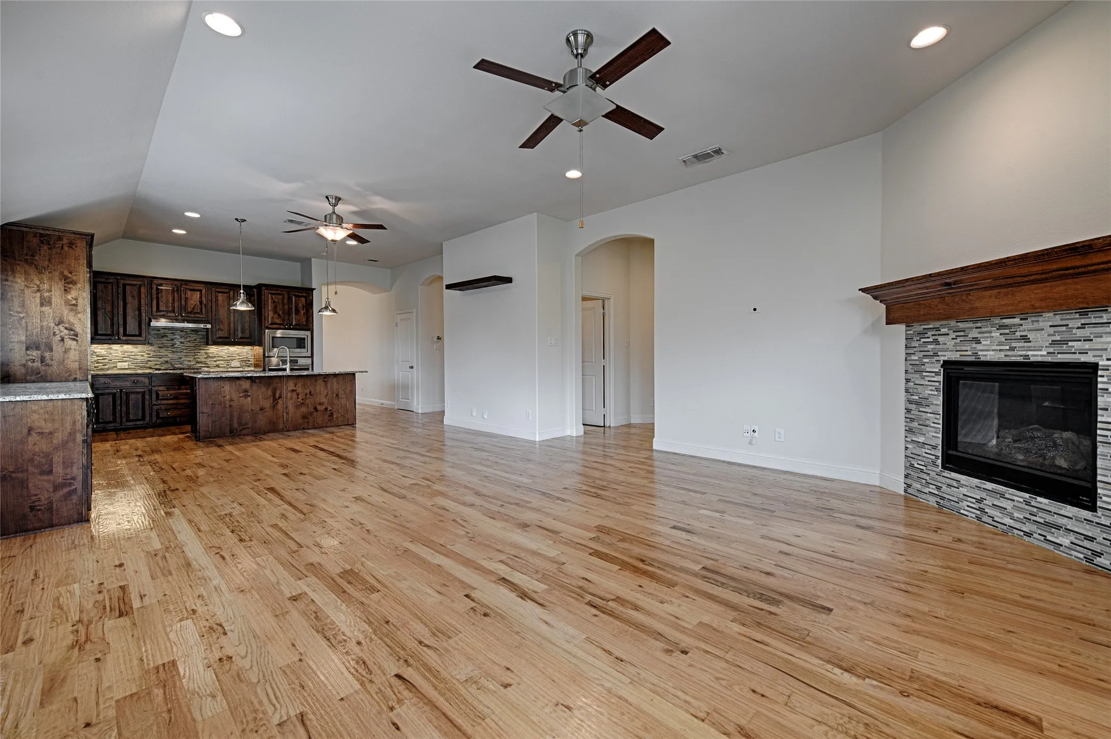 Open-concept living area featuring wood-finish flooring, a stone-clad fireplace with a wood mantle, and recessed ceiling lighting