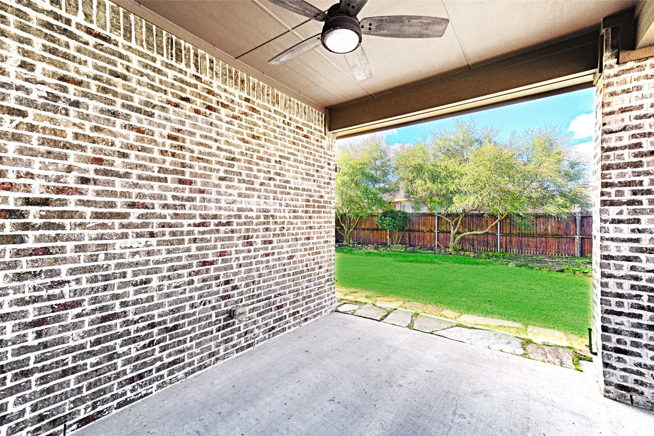 Covered concrete patio featuring a ceiling fan with integrated lighting, brick exterior walls, and a flagstone pathway