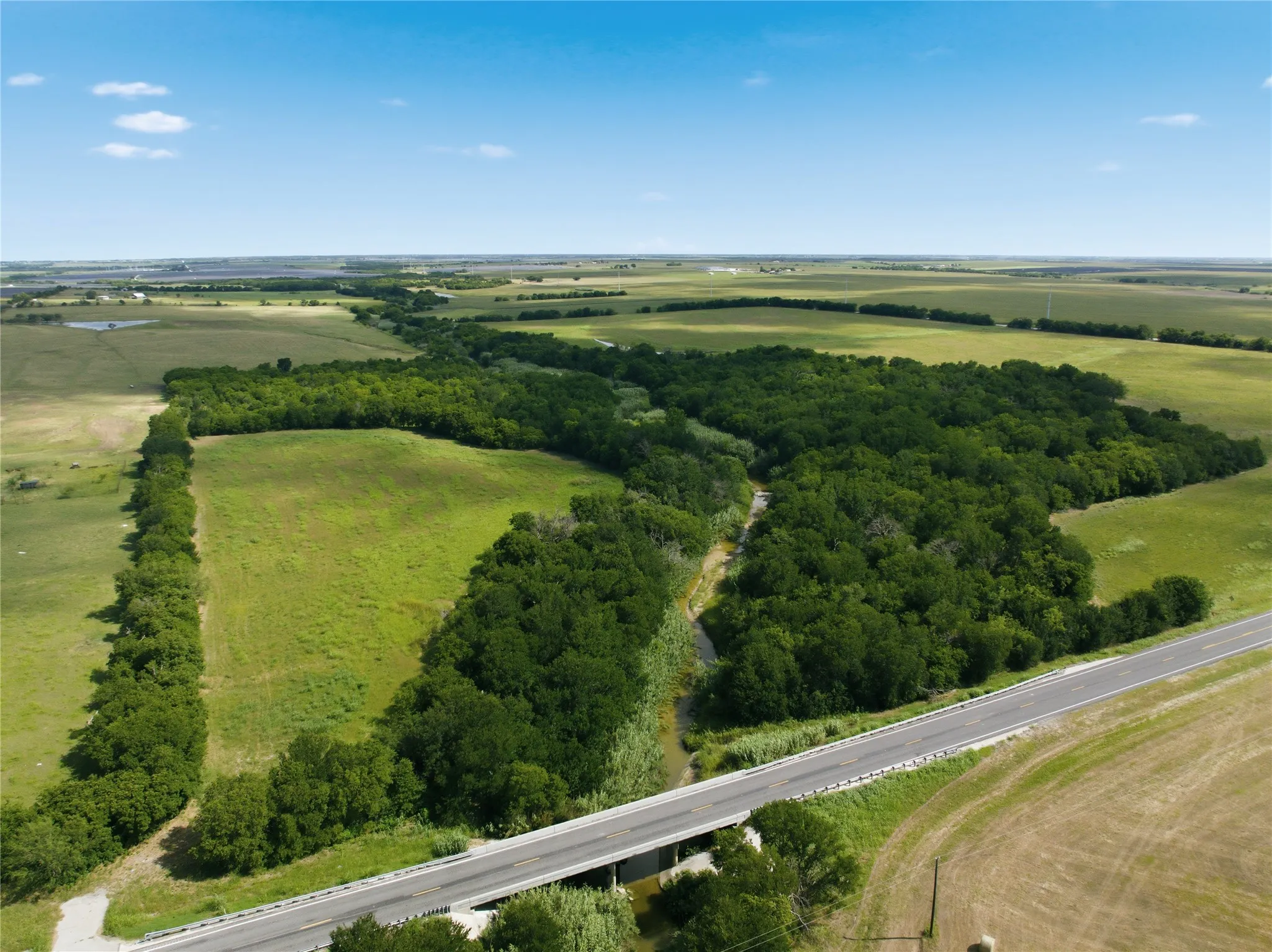 Expansive natural landscape featuring a winding waterway, mature tree lines, open green fields, and a paved road with an overpass