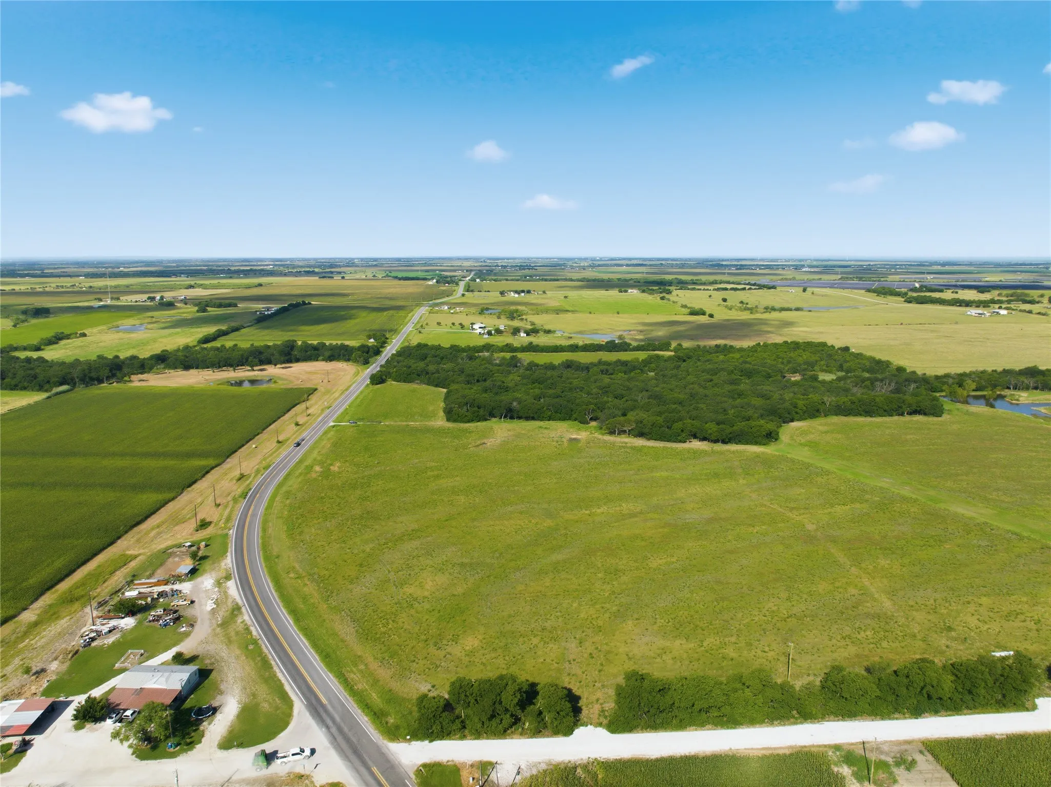 Expansive green fields featuring a clear blue sky, a paved two-lane road, scattered clusters of trees, and distant cultivated farmlands