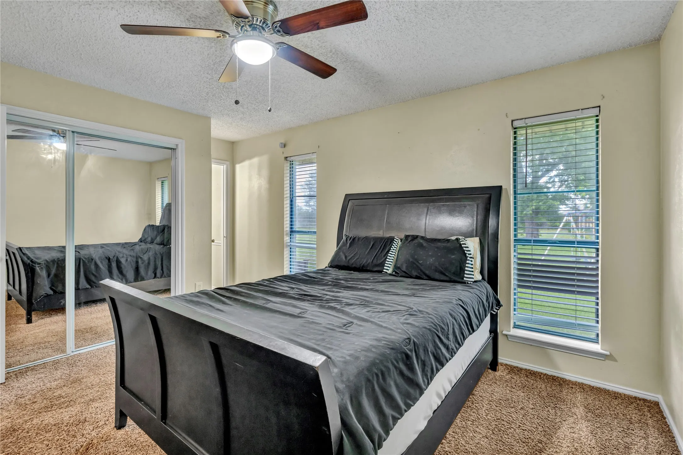 Bedroom featuring neutral wall paint, textured ceiling, brown carpeting, dual windows with blinds, and mirrored closet doors