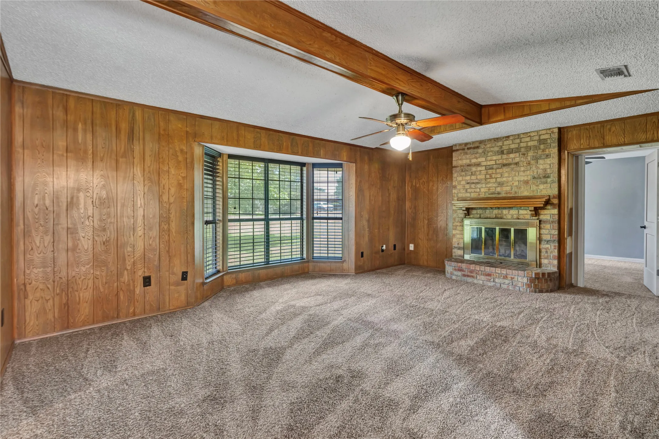 Living area featuring wood paneling, a brick fireplace with a wood mantle, a bay window with blinds, wood-finish ceiling beams, and a ceiling fan