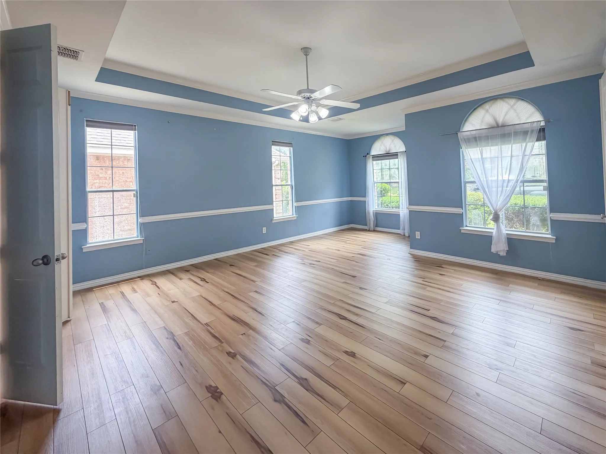 Spacious room featuring wood-finish flooring, two picture windows, two arched windows, a tray ceiling, and a ceiling fan