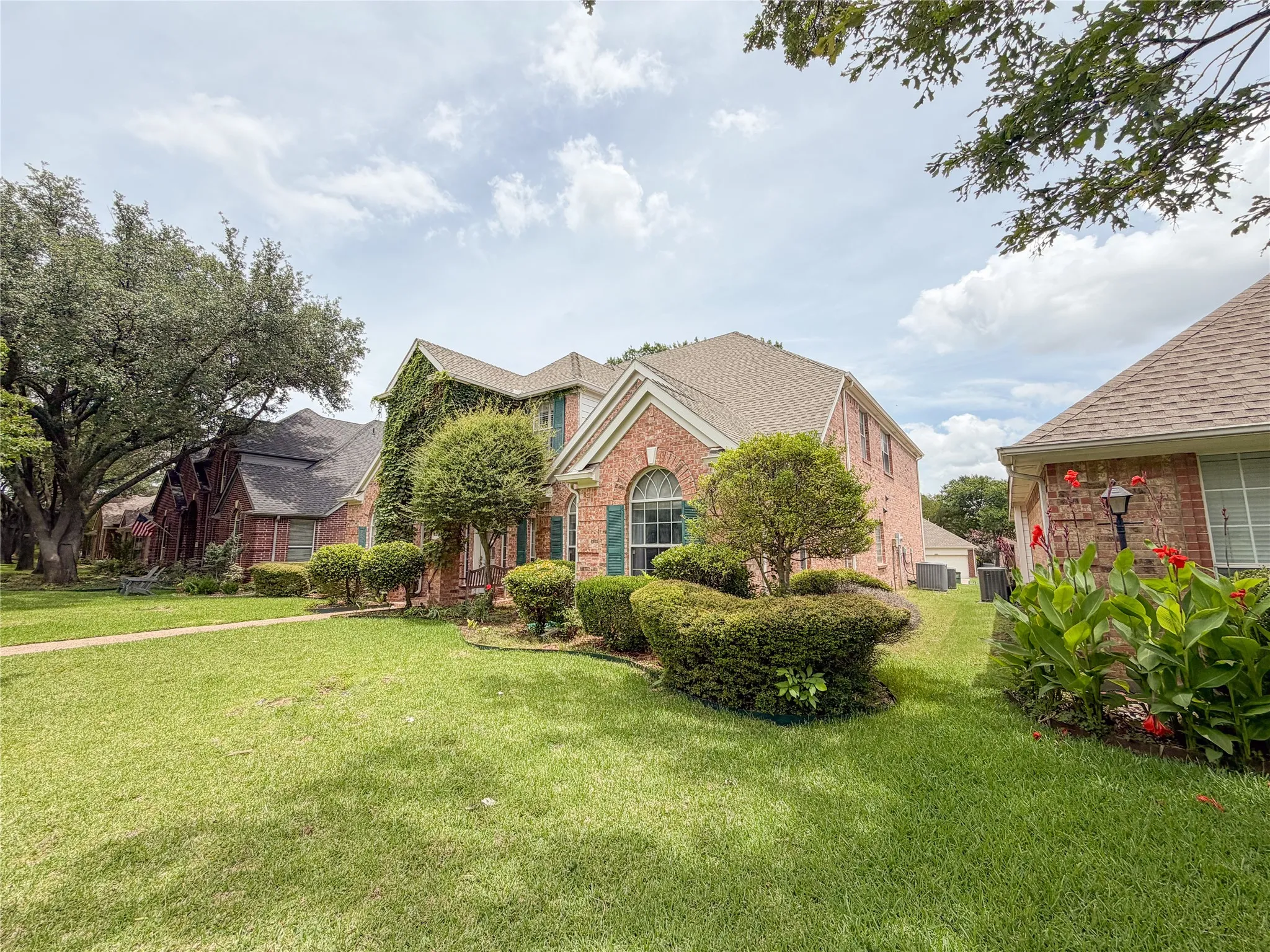 Brick exterior property featuring a gabled roof, arched window, green shutters, and established landscaping