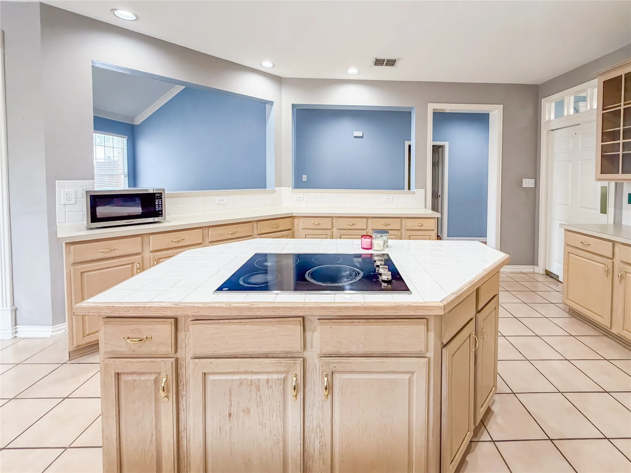 Kitchen featuring a central island with an integrated cooktop, extensive counter space with tile surfaces, light wood-finish cabinetry, and tile flooring
