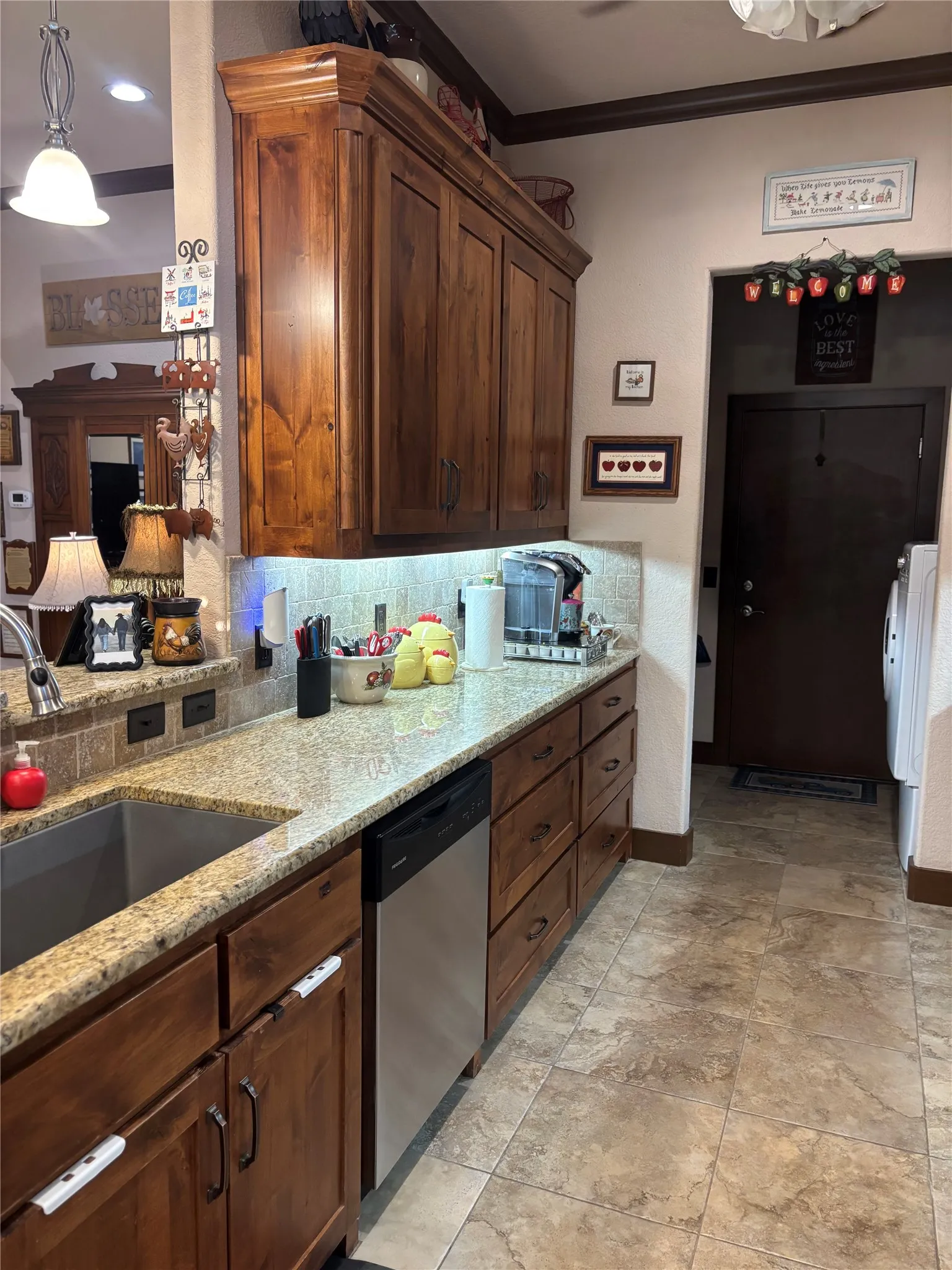 Kitchen featuring wood cabinetry with crown molding, granite countertops, stainless steel undermount sink, stainless steel dishwasher, and tile flooring