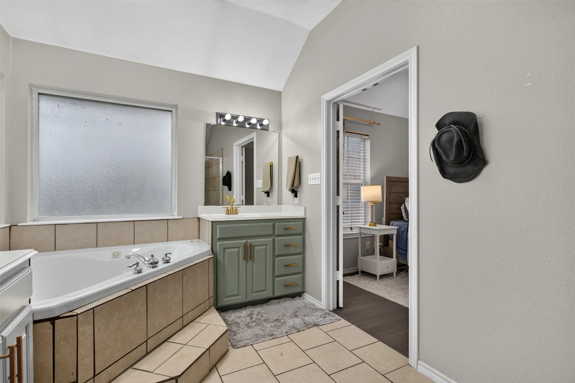 Soaking tub with tiled surround, light-toned tile flooring, dual-basin vanity with a light green cabinet finish and gold-toned hardware, and a vaulted ceiling