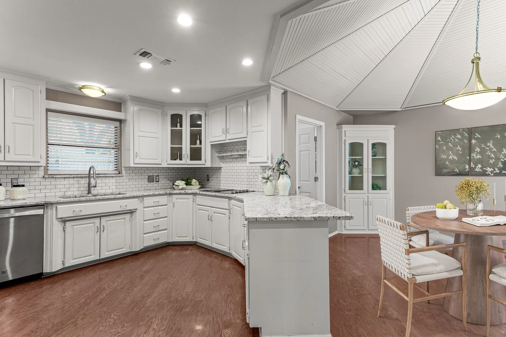 The kitchen blends timeless elegance with white cabinetry accented by silver hardware, granite countertops, double ovens, Kenmore Elite appliances, subway tile backsplash, open shelving, a pantry, butlers pantry, multiple glass-front display cabinets & a charming dining area beneath a beadboard ceiling adding character. The furniture and decor in this photo has been virtually staged, not altering the room.