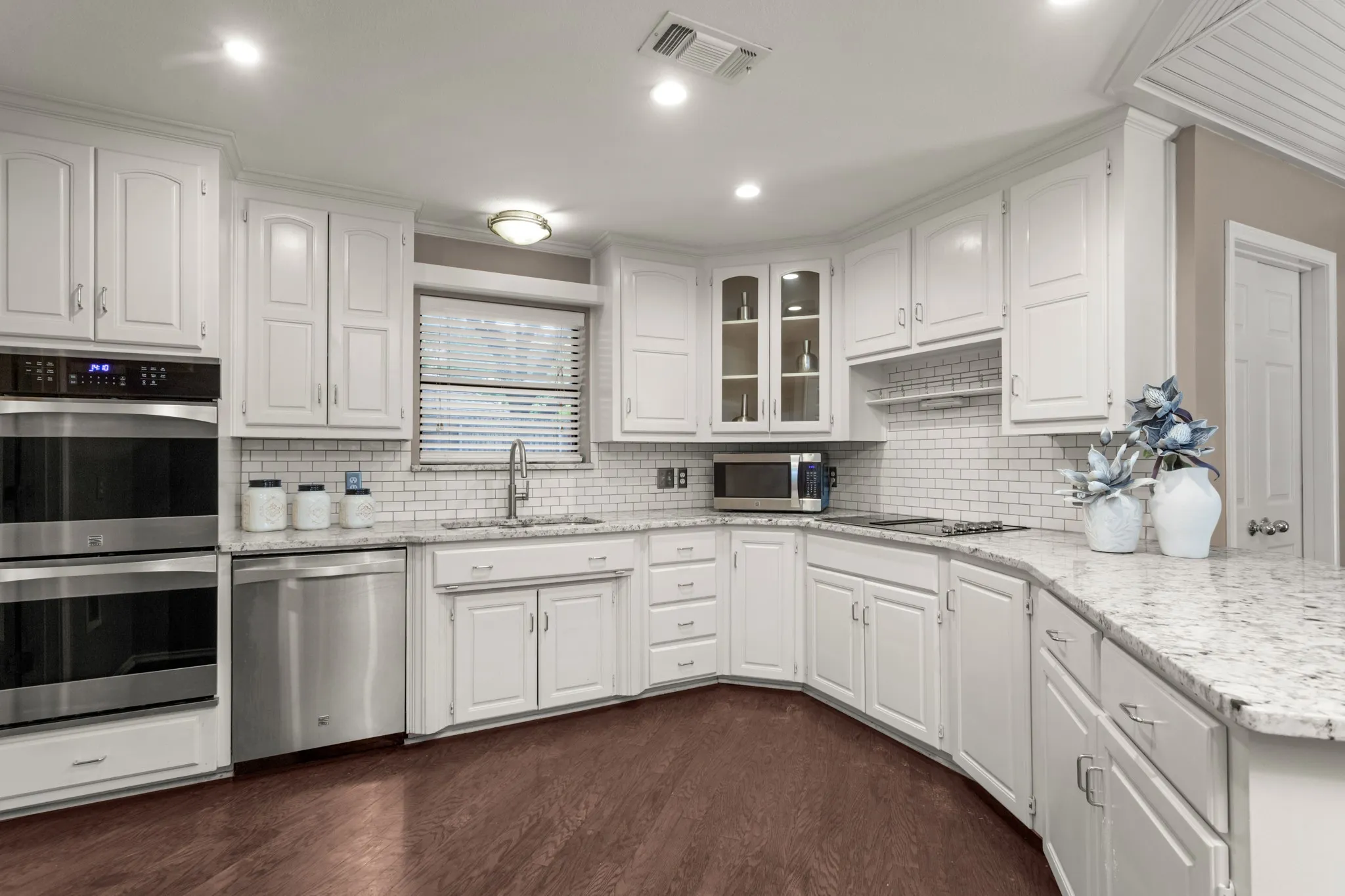 The kitchen blends timeless elegance with white cabinetry accented by silver hardware, granite countertops, double ovens, Kenmore Elite appliances, subway tile backsplash, open shelving, a pantry, butlers pantry, multiple glass-front display cabinets & a charming dining area beneath a beadboard ceiling adding character.