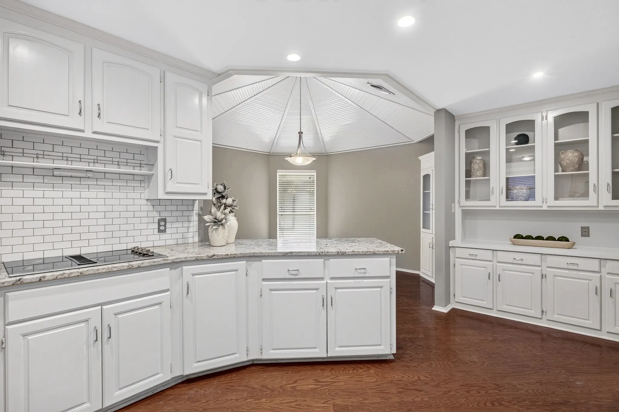 The kitchen blends timeless elegance with white cabinetry accented by silver hardware, granite countertops, double ovens, Kenmore Elite appliances, subway tile backsplash, open shelving, a pantry, butlers pantry, multiple glass-front display cabinets & a charming dining area beneath a beadboard ceiling adding character.
