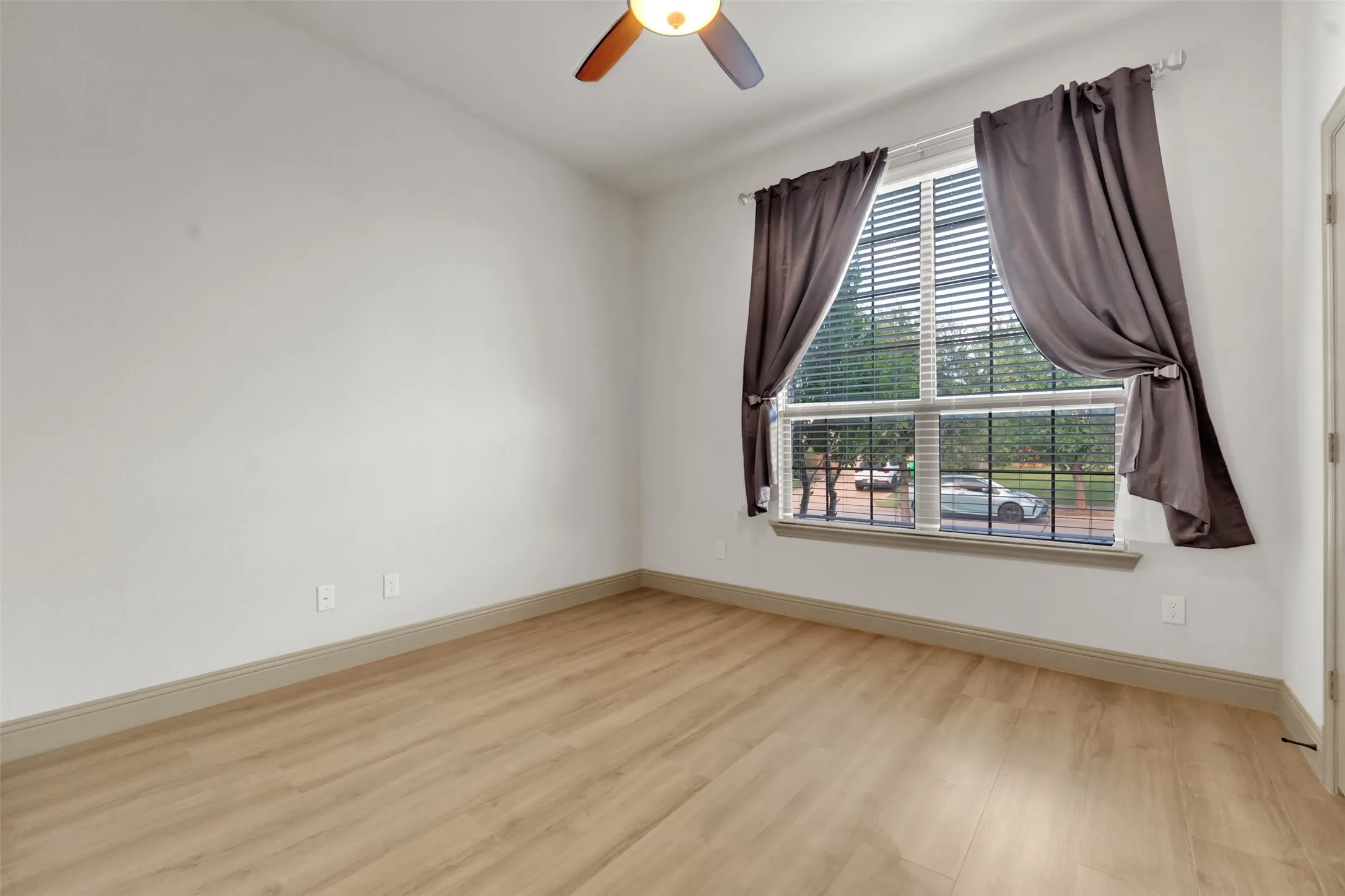 Room featuring wood-finish flooring, a ceiling fan with integrated lighting, and a large window with blinds and dark drapery
