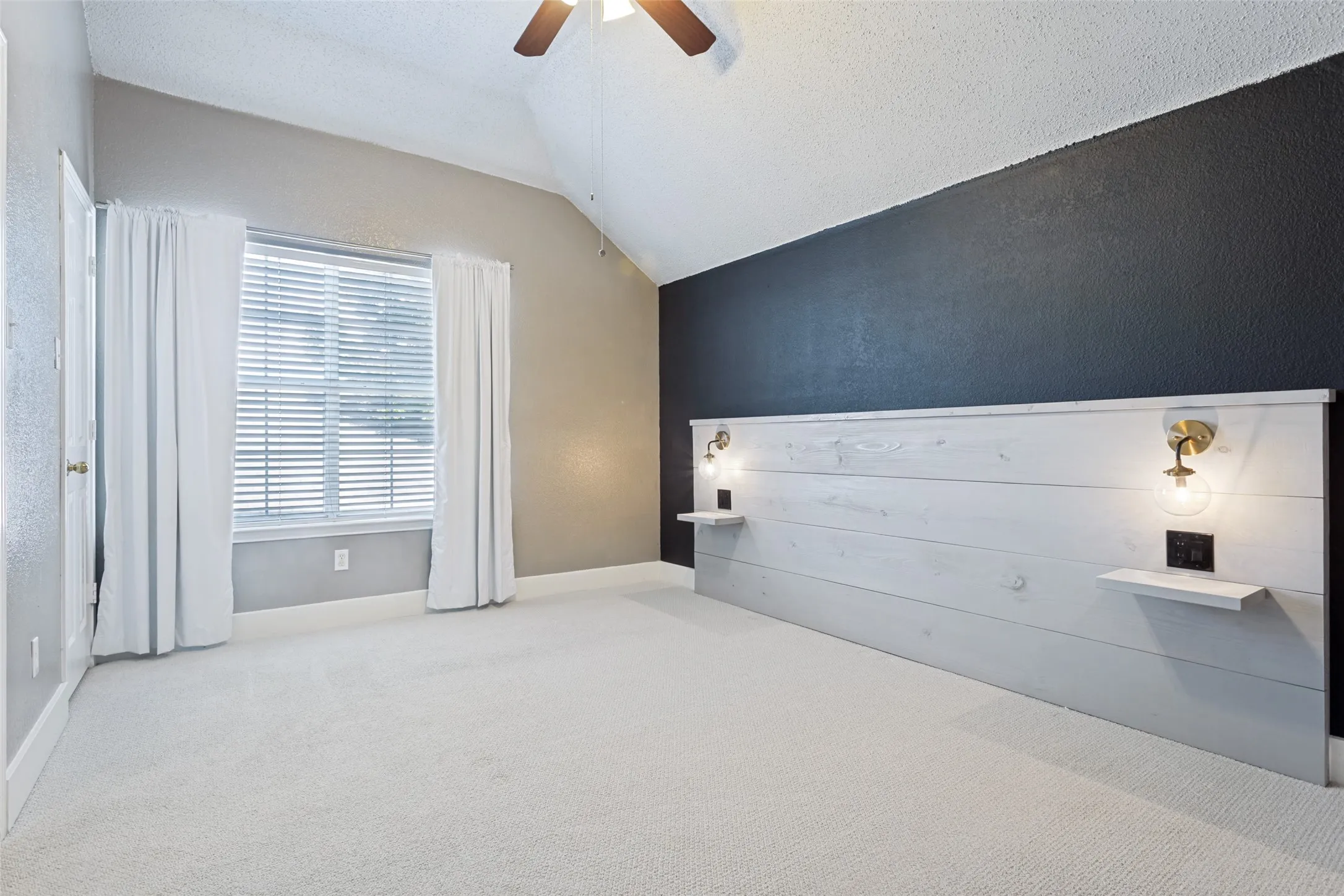 Vaulted ceiling with a ceiling fan, light-colored carpet, a window with blinds, and an accent wall featuring a built-in headboard with integrated lighting and shelves