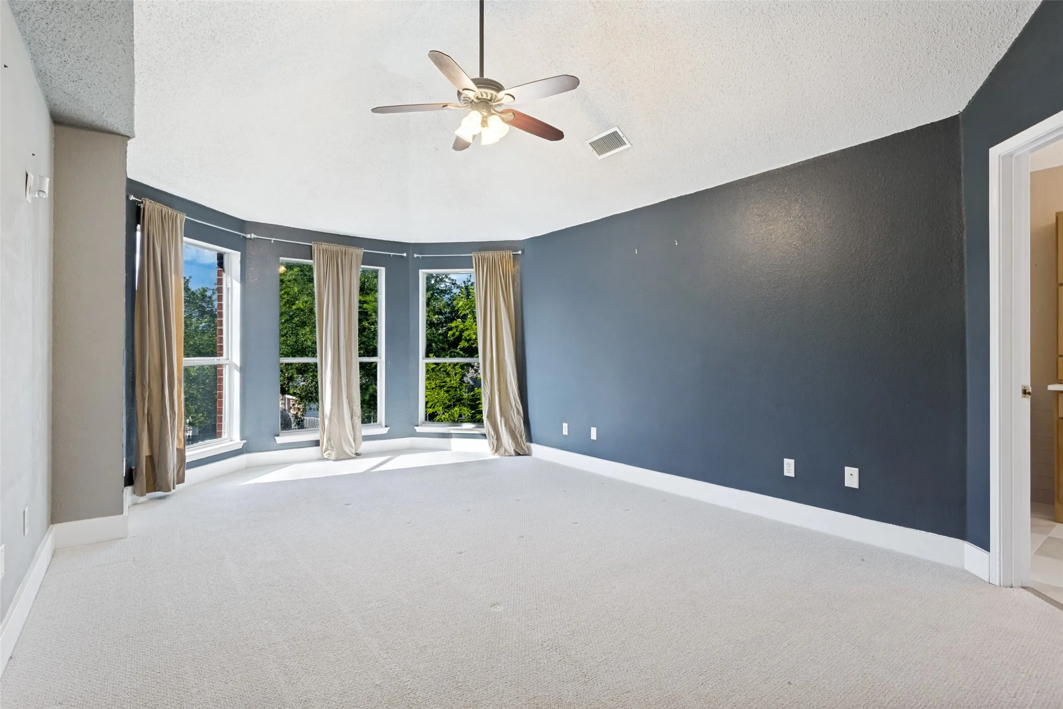 Spacious room featuring a curved wall with three large windows, light-toned carpeting, a ceiling fan, and a neutral wall palette