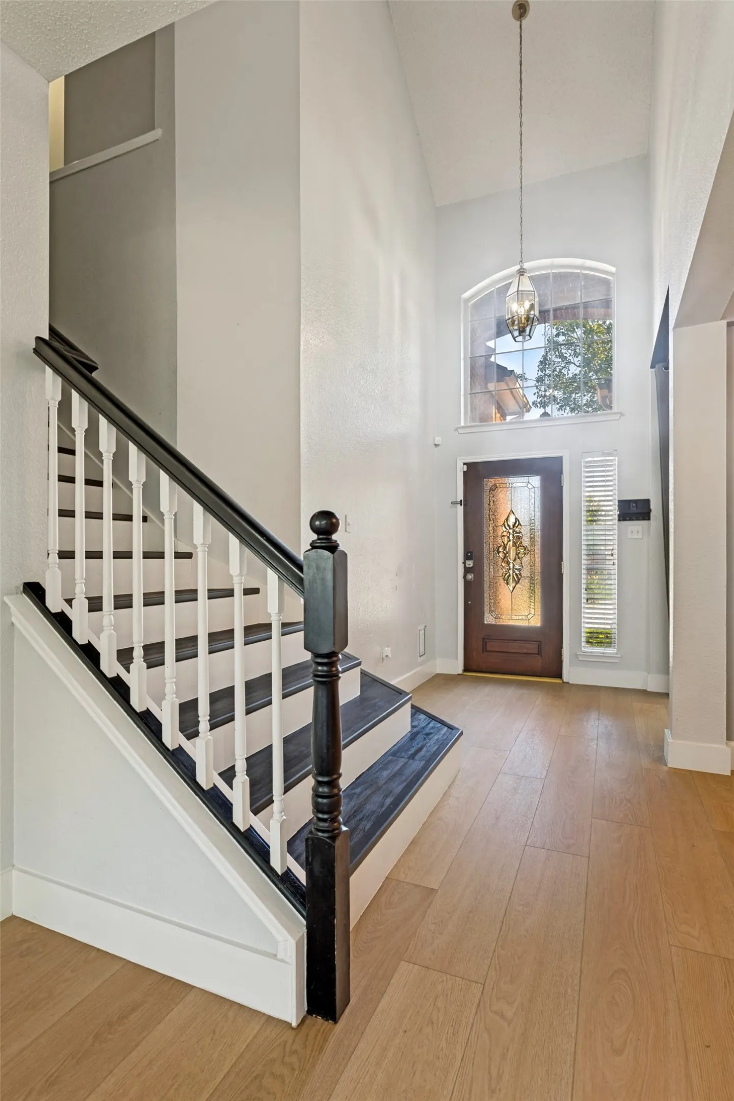 Two-story foyer featuring a staircase with white balusters and dark wood-finish treads, light wood-finish flooring, a decorative front door with ornate glass, an arched transom window, and a vaulted ceiling with a pendant light fixture