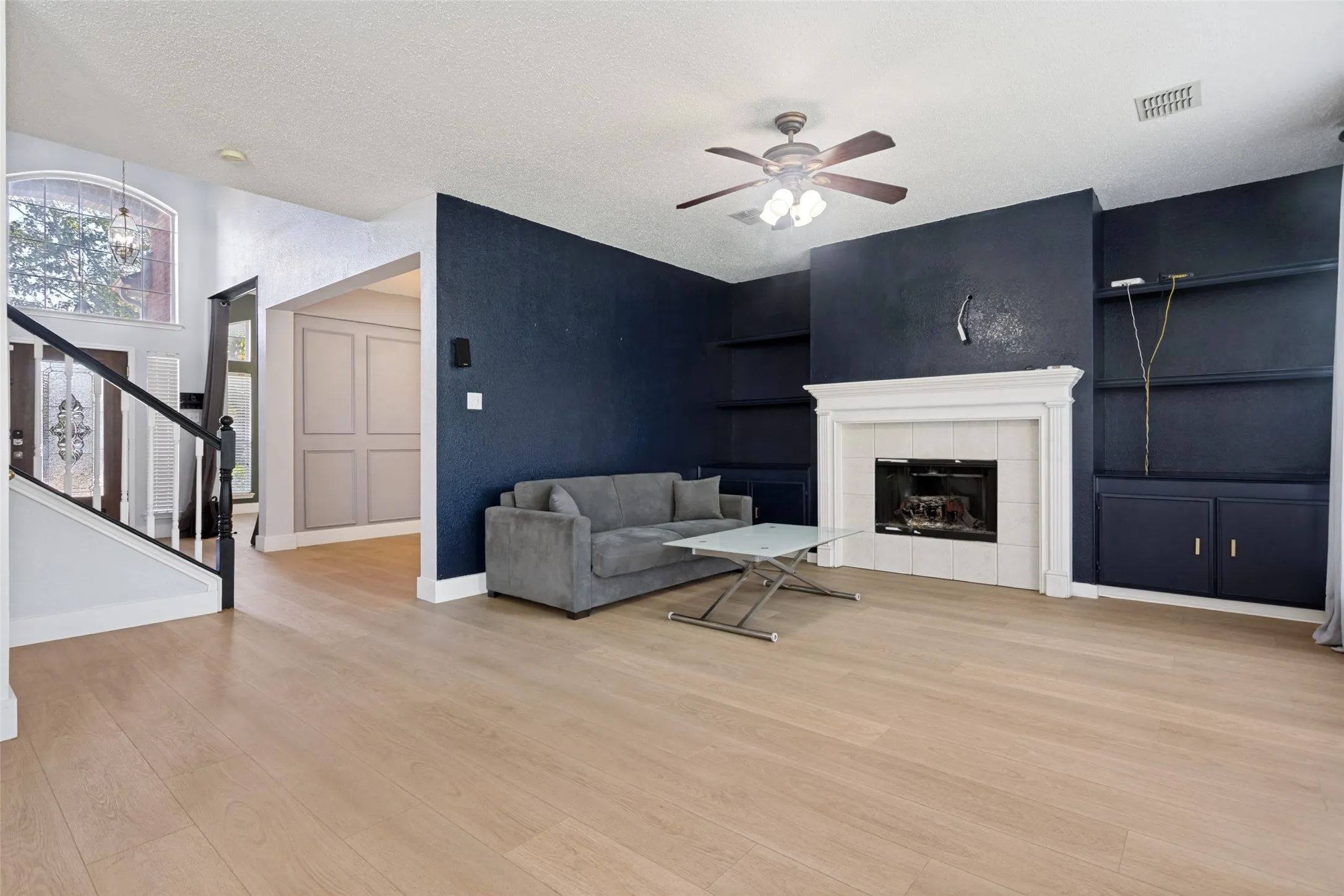 Spacious living area featuring light wood-finish flooring, a white-mantled fireplace with tile surround, built-in shelving, and a ceiling fan