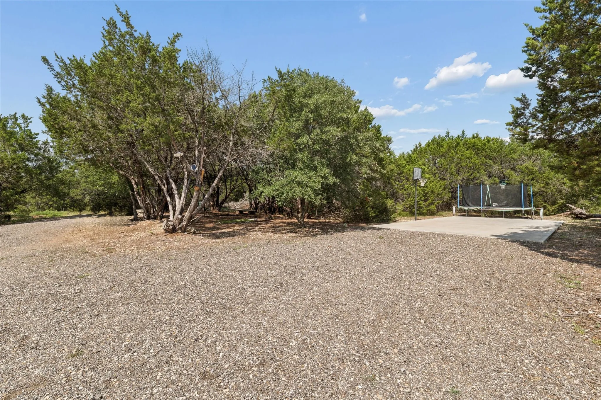 Expansive gravel area leading to a concrete pad, surrounded by mature trees