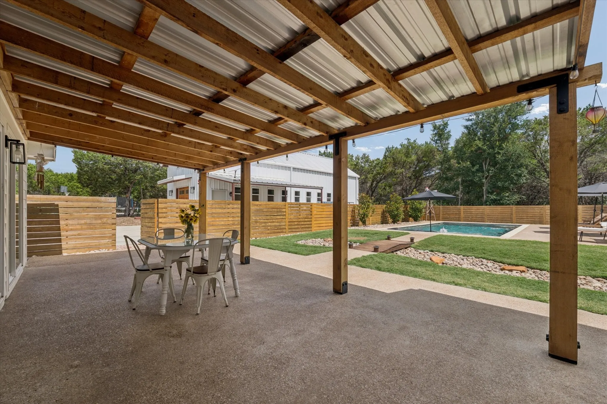Expansive outdoor living area featuring a covered patio with a wood-framed metal roof, concrete flooring, and an in-ground swimming pool