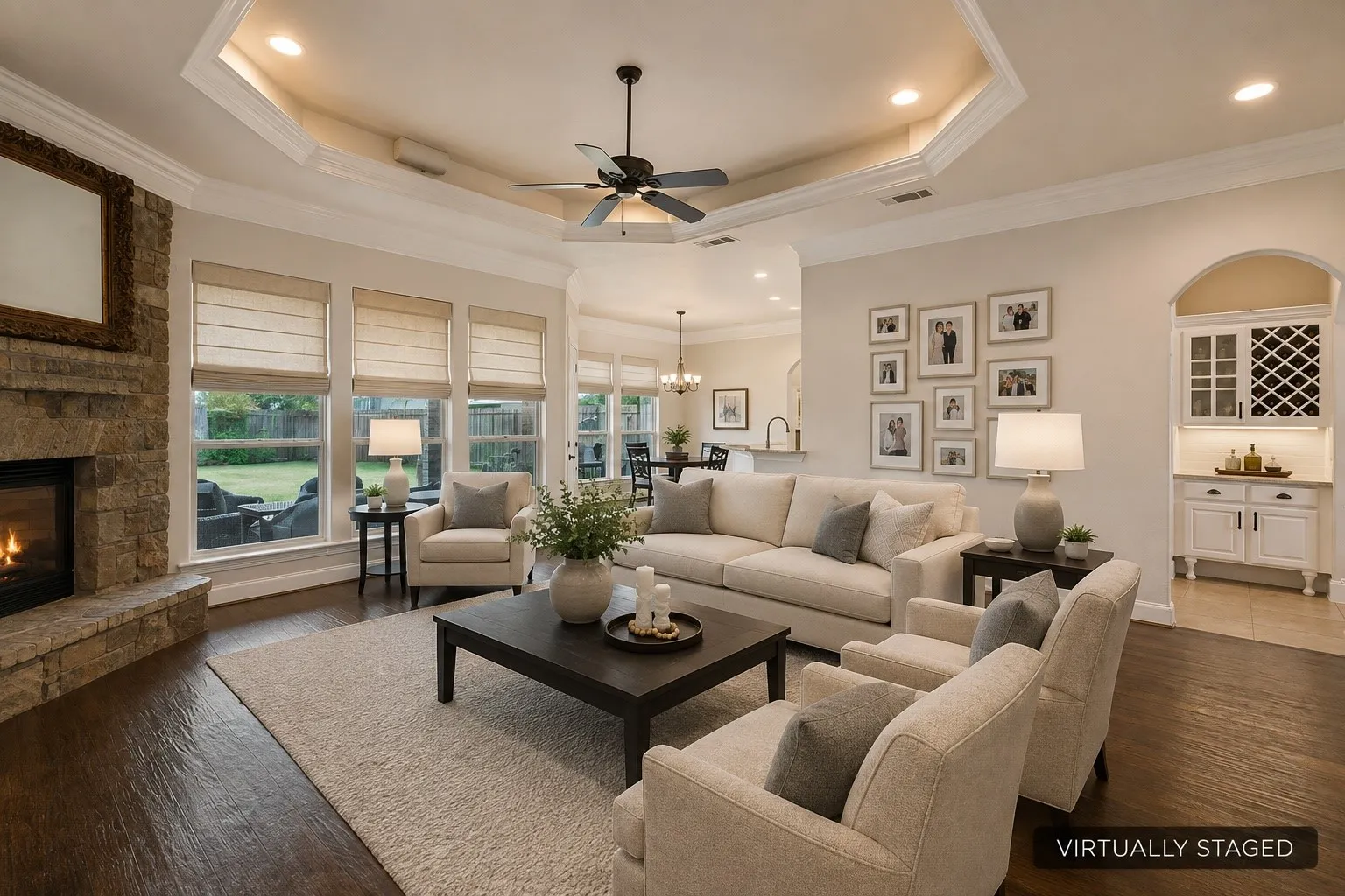 Stone-faced fireplace, coffered ceiling with recessed lighting, dark wood-finish flooring, built-in wet bar with cabinetry, and multiple windows with roller shades