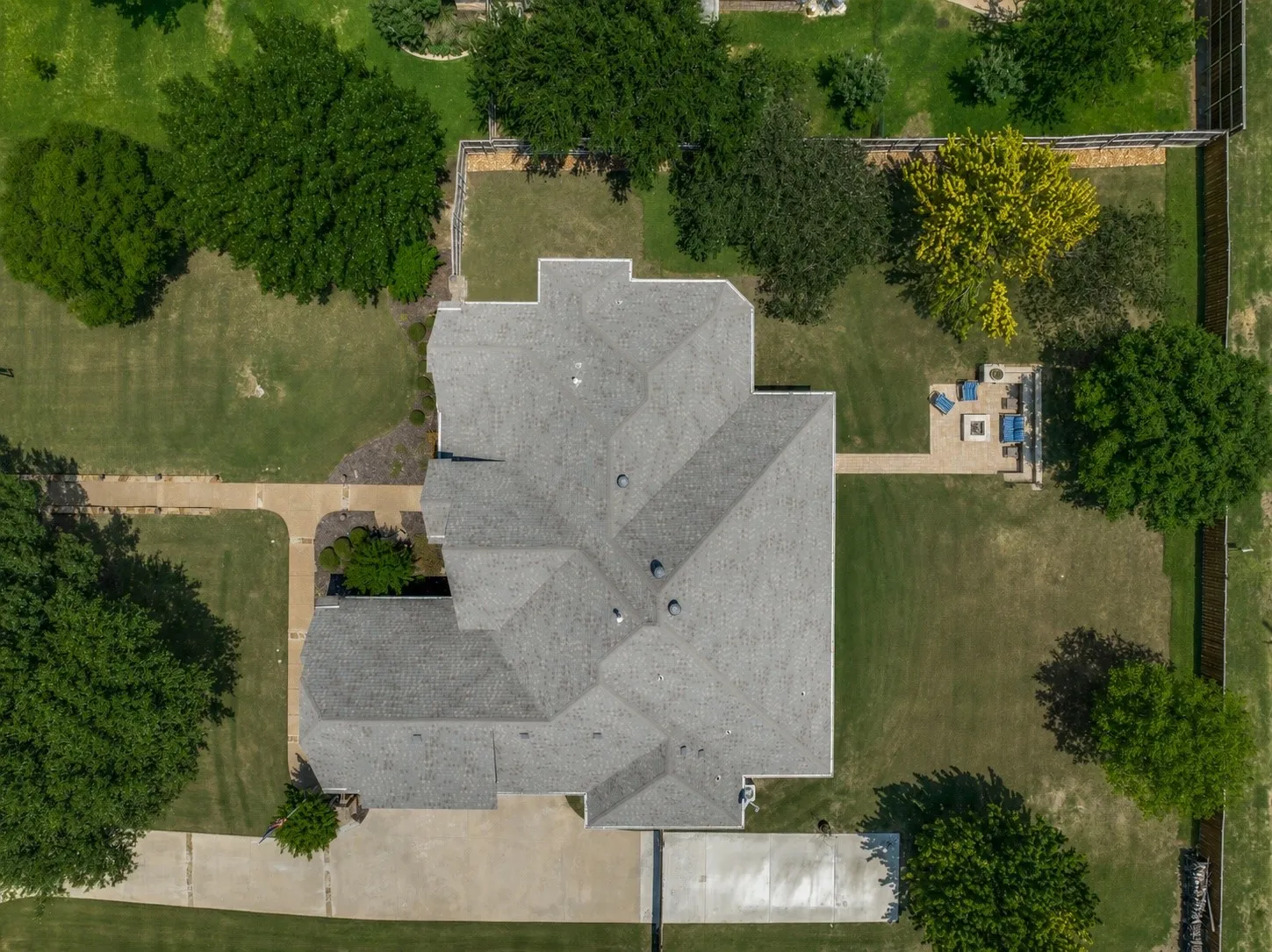 Expansive property featuring a grey shingle roof, paved driveway, multiple mature trees, and a backyard patio with a built-in fire pit