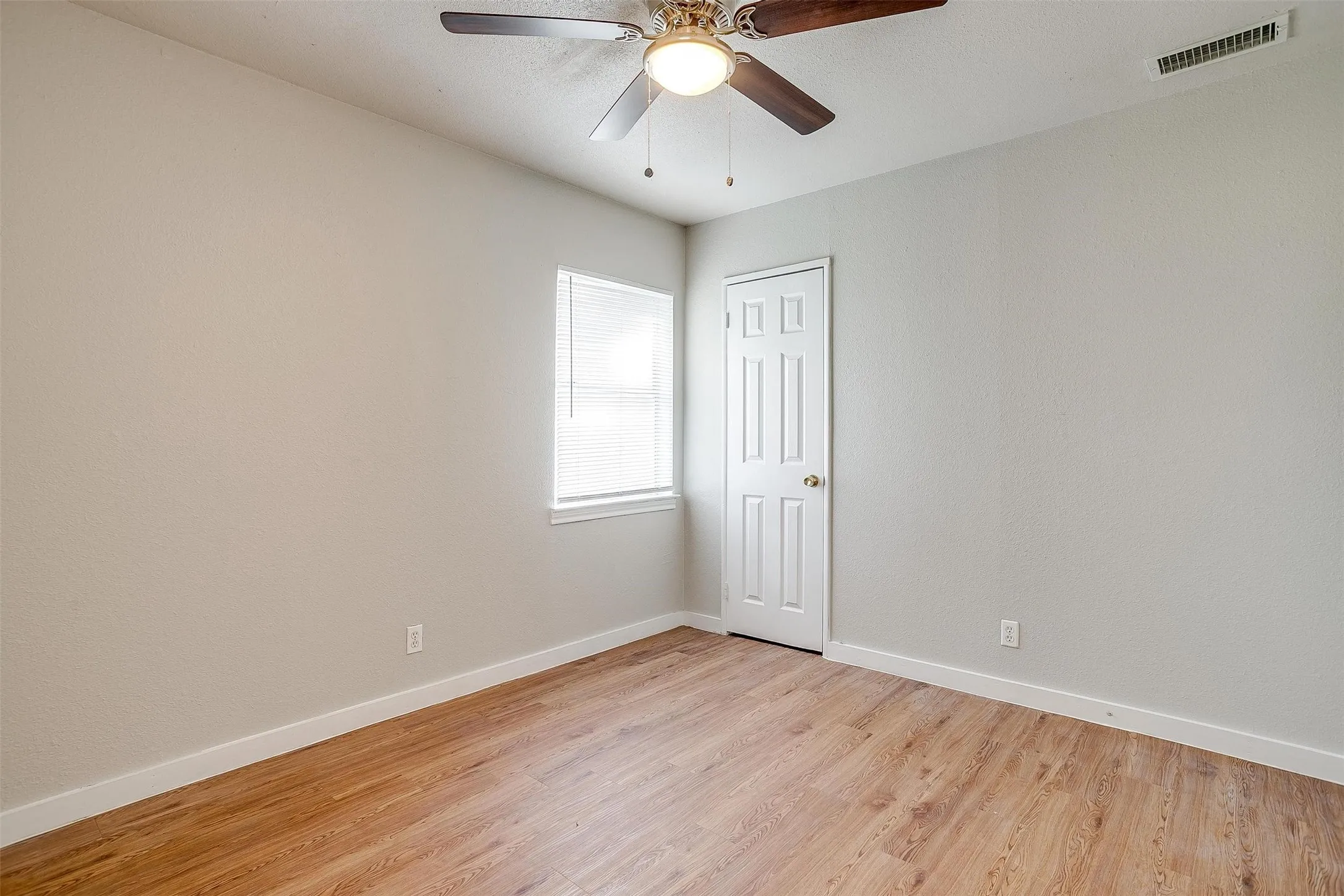 Spacious room featuring wood-finish flooring, light neutral-toned walls, a ceiling fan with integrated lighting, a paneled door, and a window with blinds