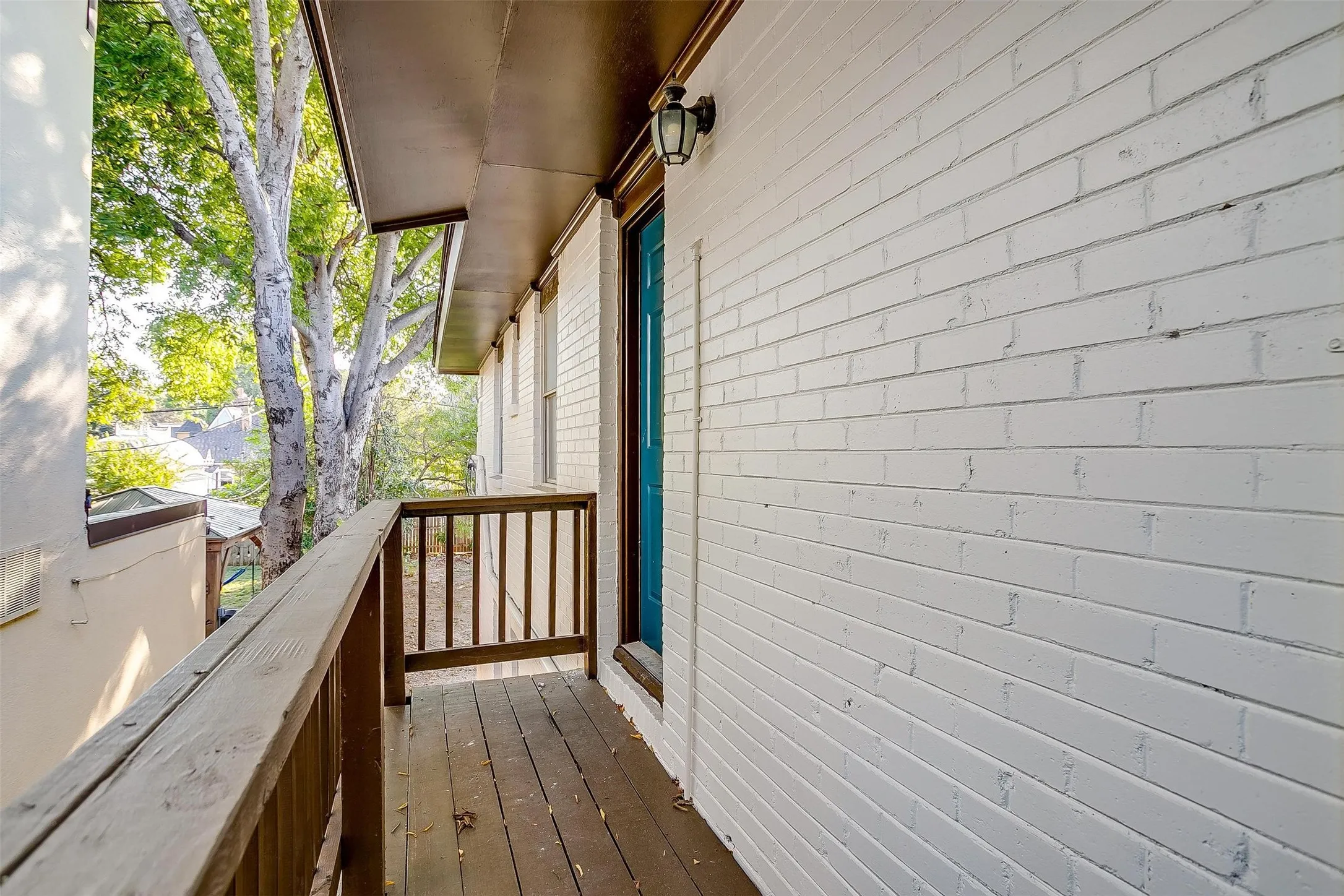 Outdoor wooden balcony with a wood railing system, white painted brick exterior, and a teal entry door