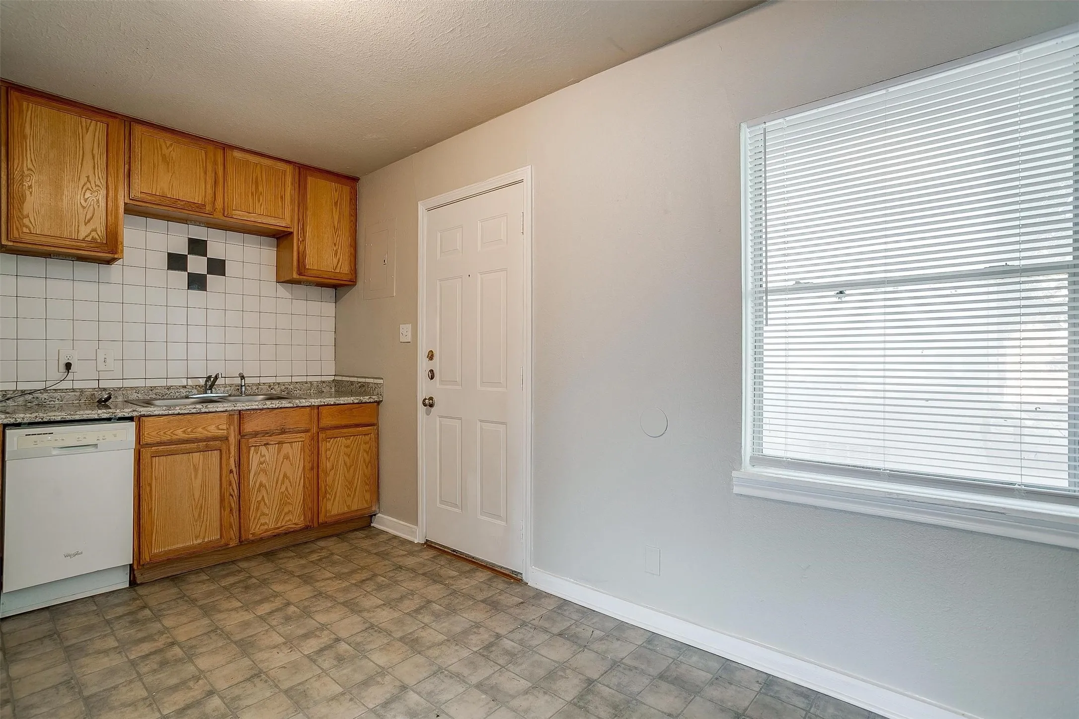 Kitchen featuring wood cabinetry, a white tile backsplash with decorative black accents, granite-style countertops, a white dishwasher, and durable flooring