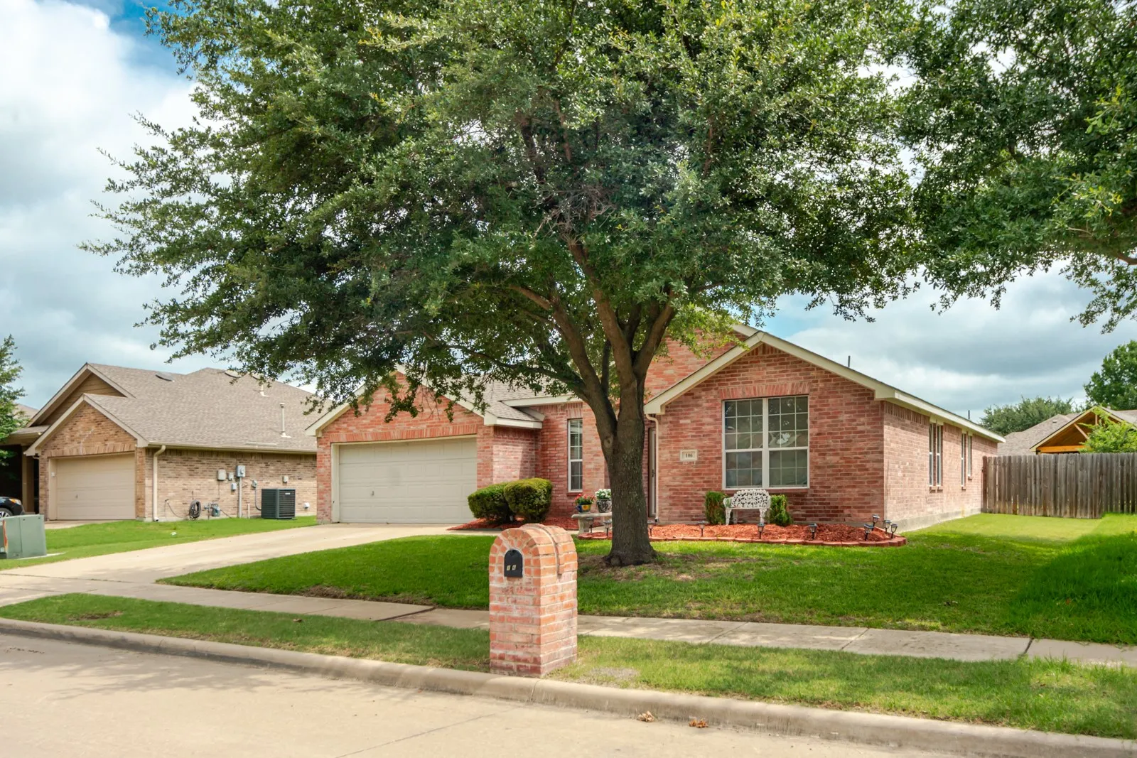 Brick exterior featuring a two-car garage, mature trees, a manicured lawn, and a brick mailbox