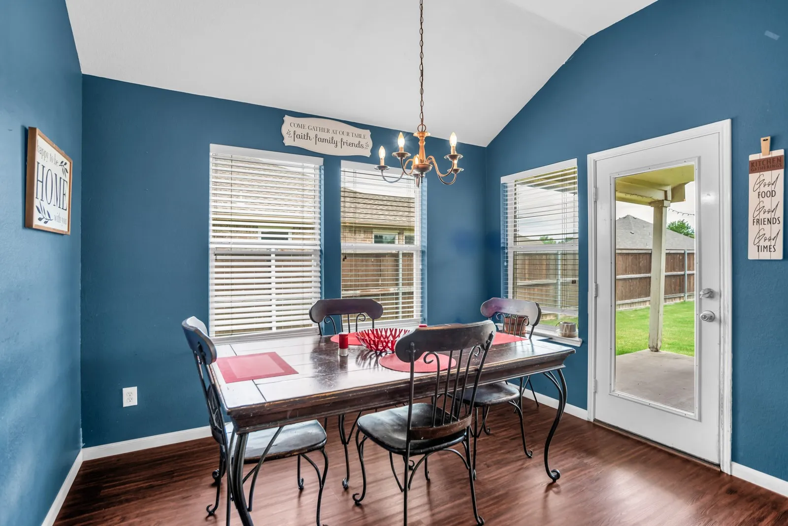 Vaulted ceiling dining area featuring wood-finish flooring, multiple windows with blinds, a glass-panel exterior door, and a bronze-finish chandelier