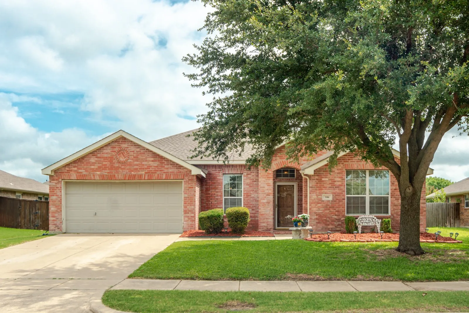 Traditional brick exterior residence featuring an attached two-car garage, mature front yard tree, arched entryway, and a concrete driveway