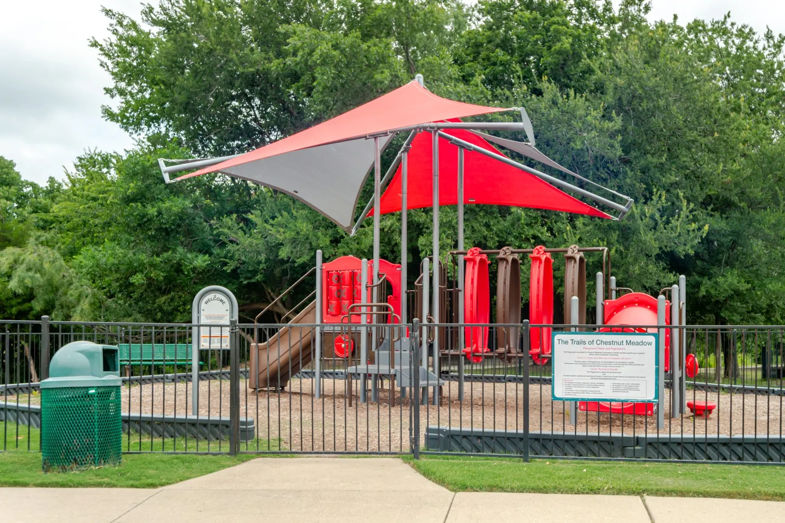 Fenced community playground featuring a multi-level play structure, slides, and red shade canopies