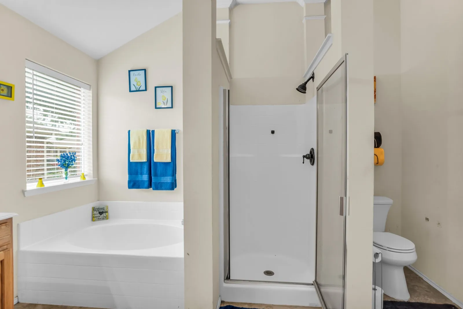 Bathroom with a built-in corner soaking tub featuring a window with blinds, a glass-enclosed shower with dark fixtures, and a commode