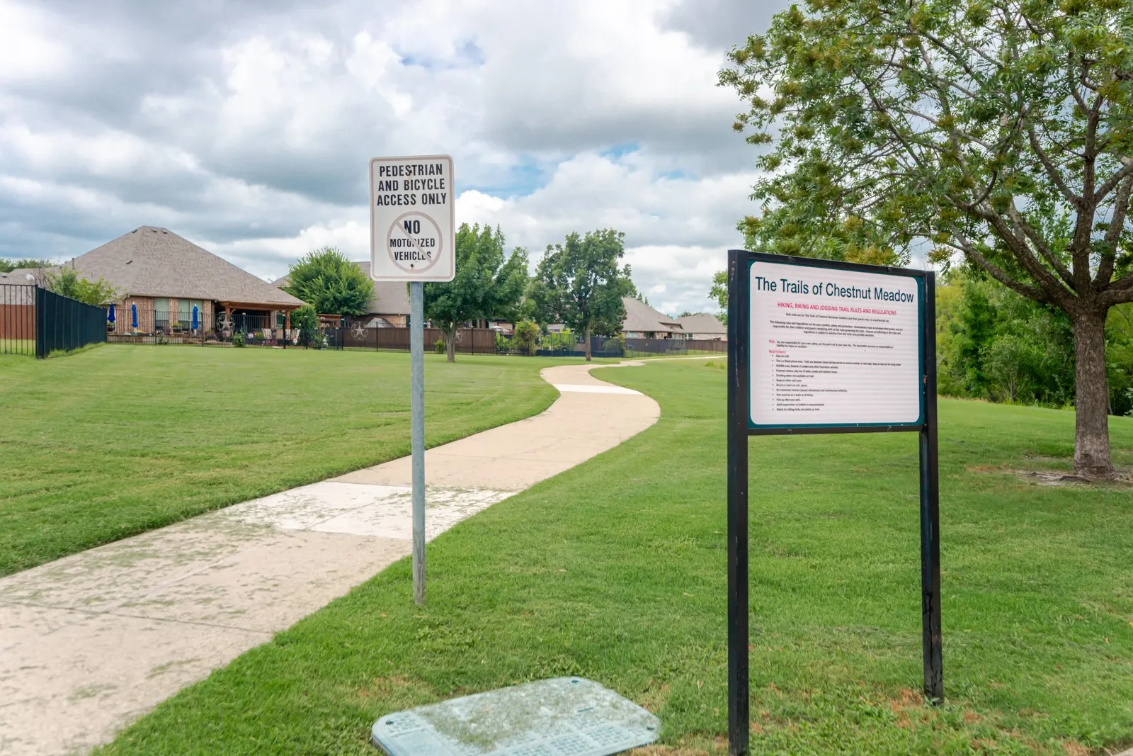 Paved walking path bordered by expansive green lawns and mature trees, featuring a community informational sign