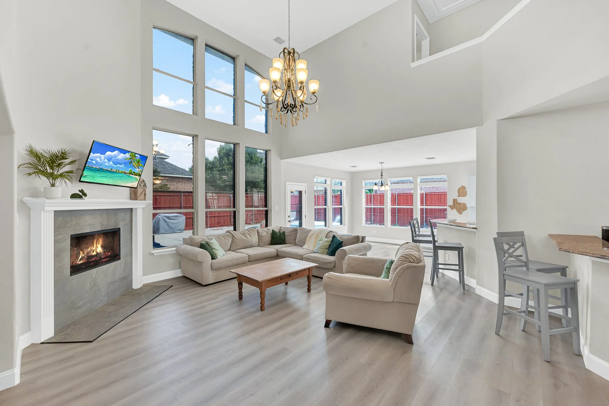 Soaring ceilings with a decorative chandelier, large windows, a tiled fireplace with a white mantel, wood-finish flooring, and a breakfast bar with a stone-look countertop
