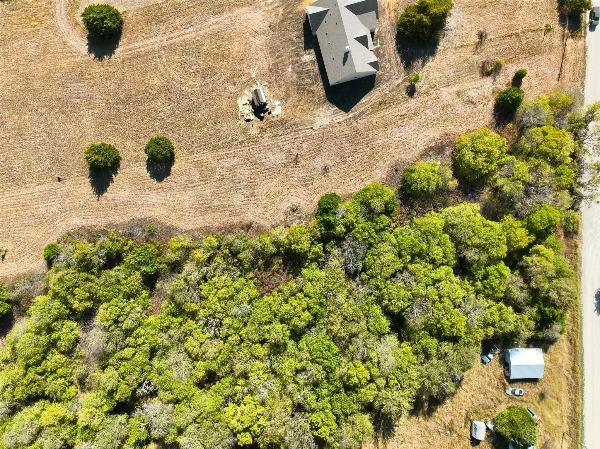Aerial perspective showcasing a parcel of land featuring a single-family residence with a dark-pitched roof, mature trees, dense green foliage, and a gravel road