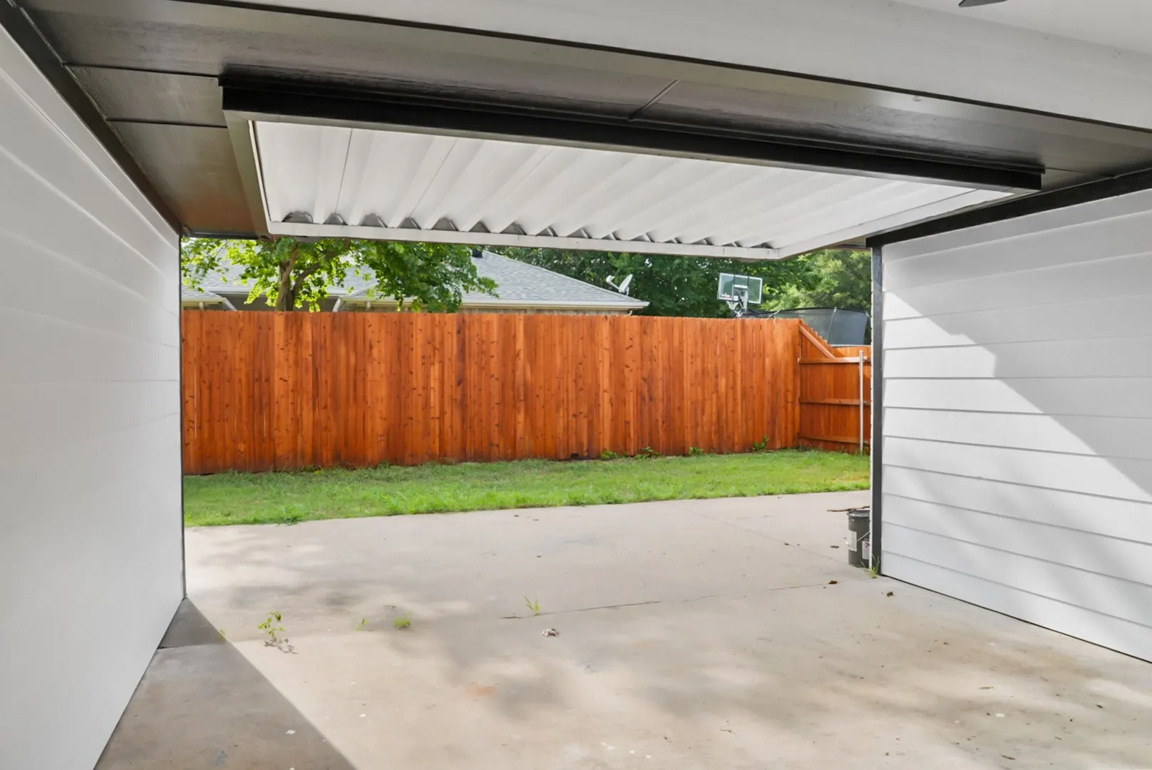 Covered patio with concrete flooring, framed by a newly stained wood fence and a green lawn