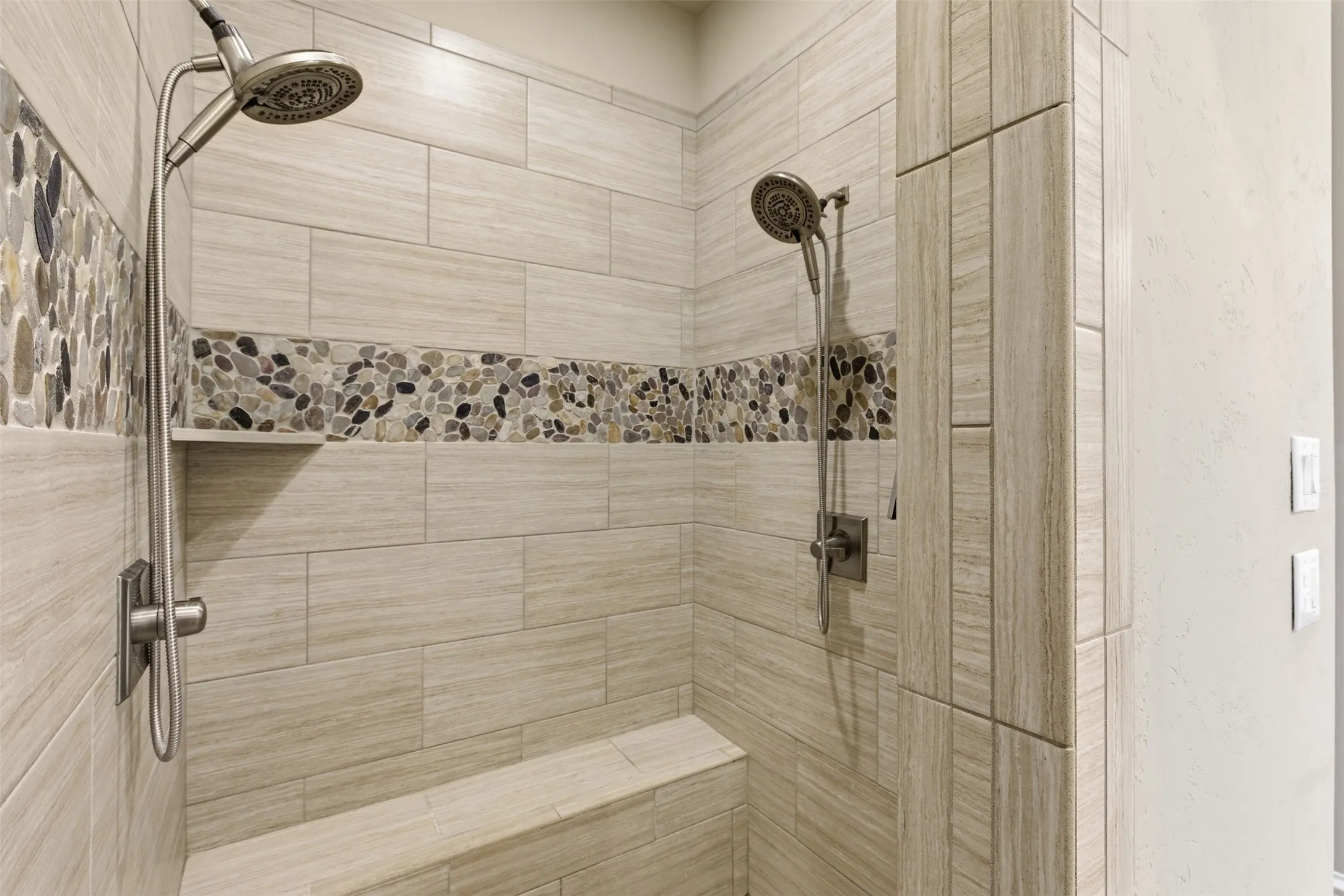 Walk-in shower featuring dual shower heads, a built-in bench, a recessed niche, and a decorative pebble tile accent band