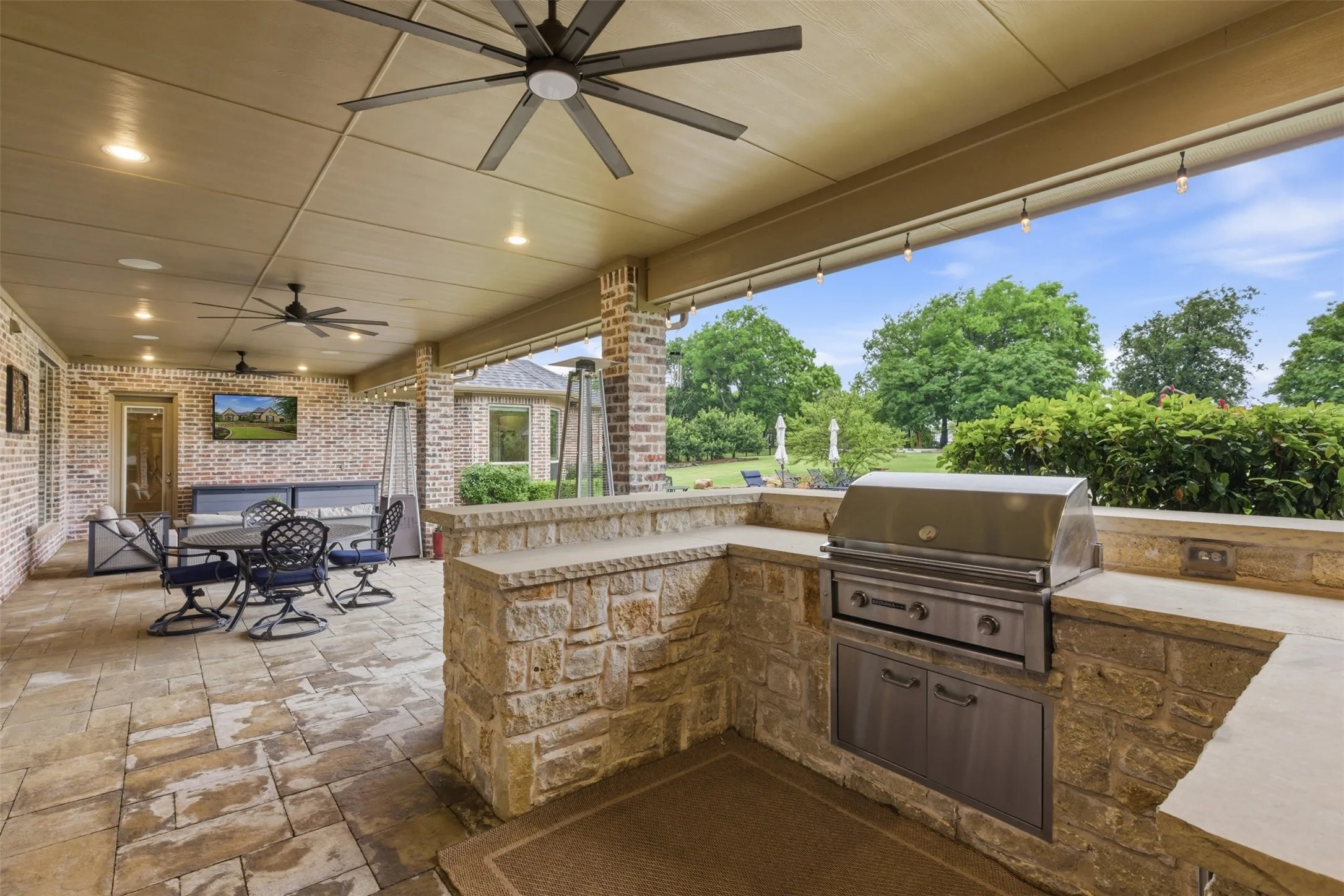 Expansive covered patio featuring a built-in stone outdoor kitchen with a stainless steel grill, recessed lighting, and multiple ceiling fans