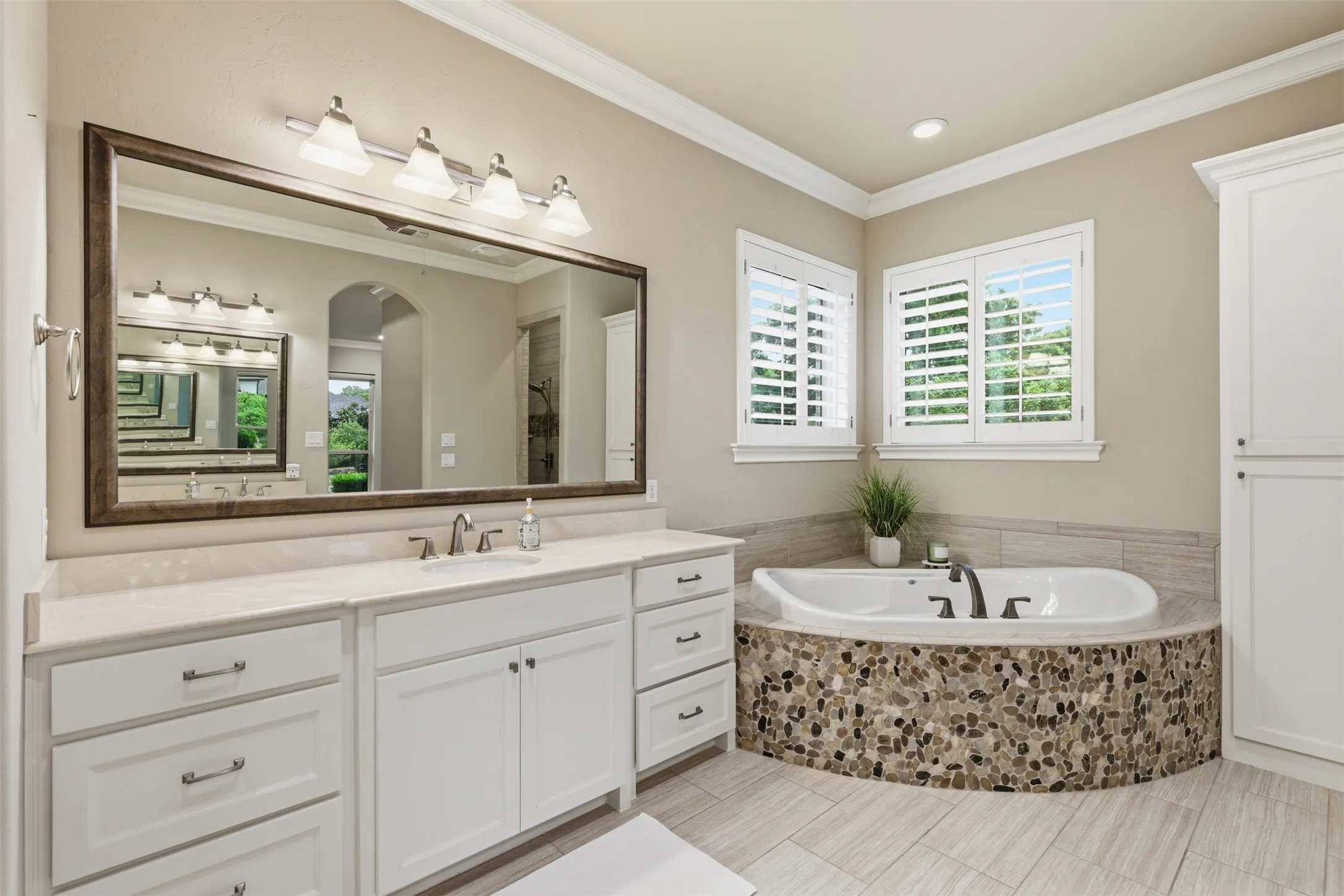 Spacious bathroom featuring a built-in tub with river rock surround, dual vanity with undermount sinks, and plantation shutters